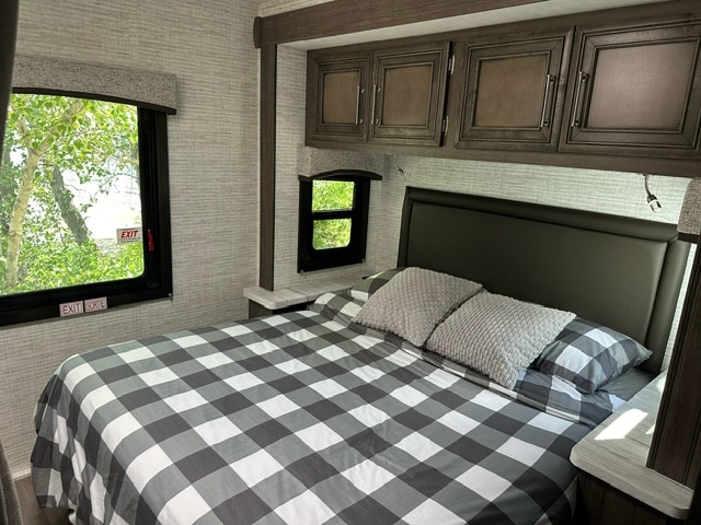 Cozy RV rental bedroom featuring a plaid bed, overhead cabinets, and windows showing green trees outside.. Entegra Coach Odyssey SE 22C 2024