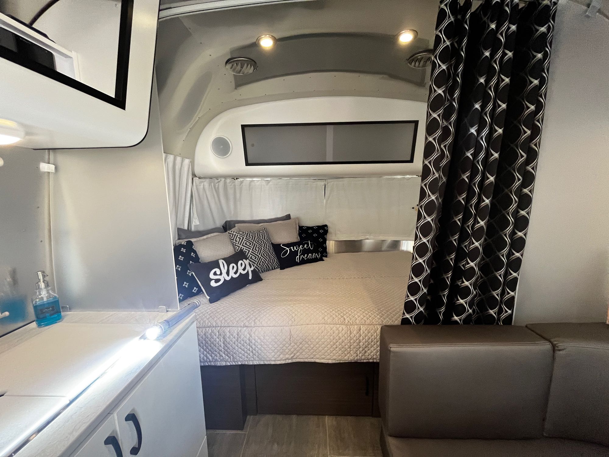 This RV rental shows a cozy bedroom with a quilted bed, decorative pillows, and a stylish black curtain, ideal for a comfortable stay.. Airstream Caravel 2021