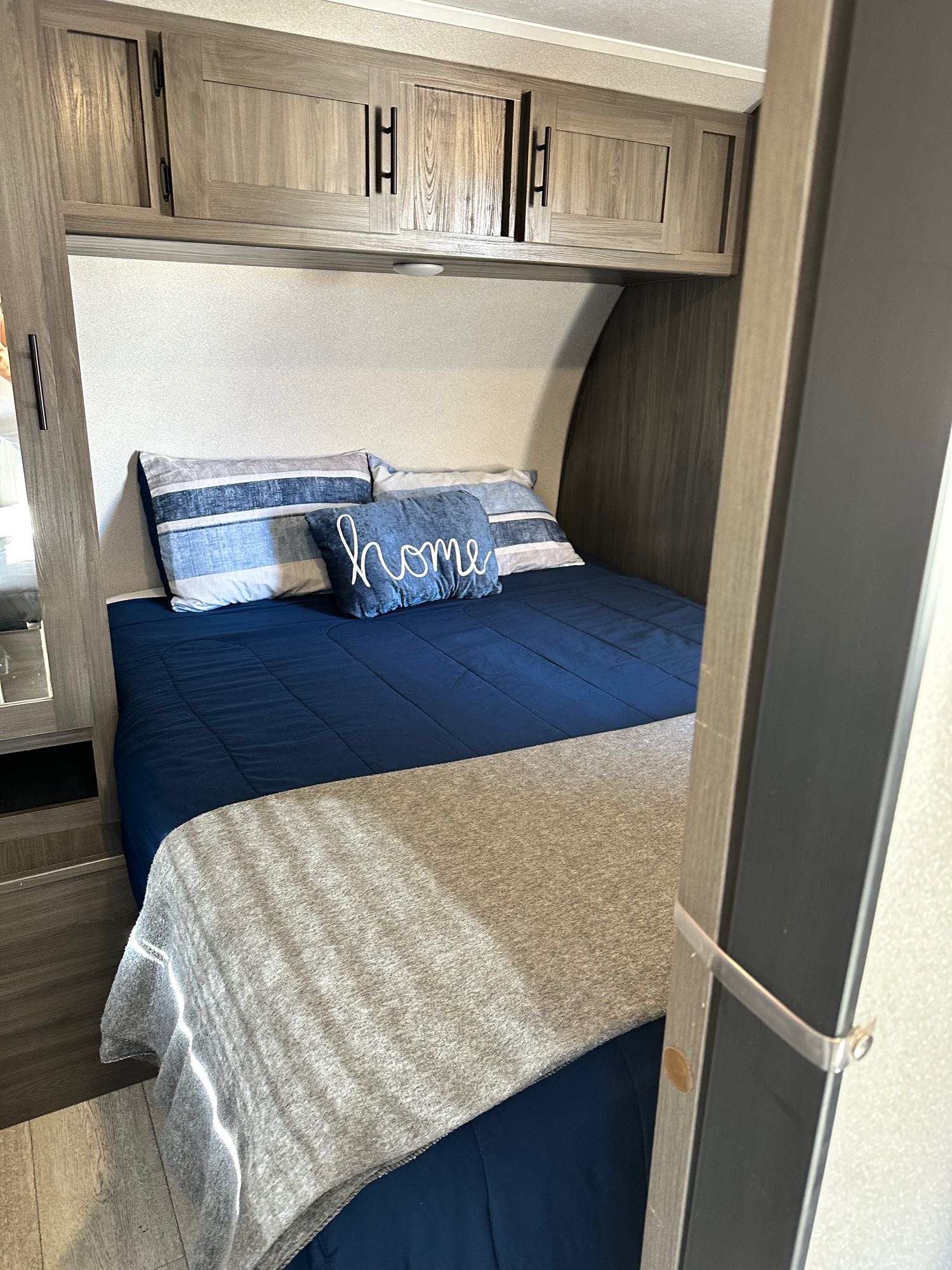 Explore a cozy RV rental bedroom featuring a blue bed, striped pillows, and ample overhead storage cabinets.. Shasta 26db 2021