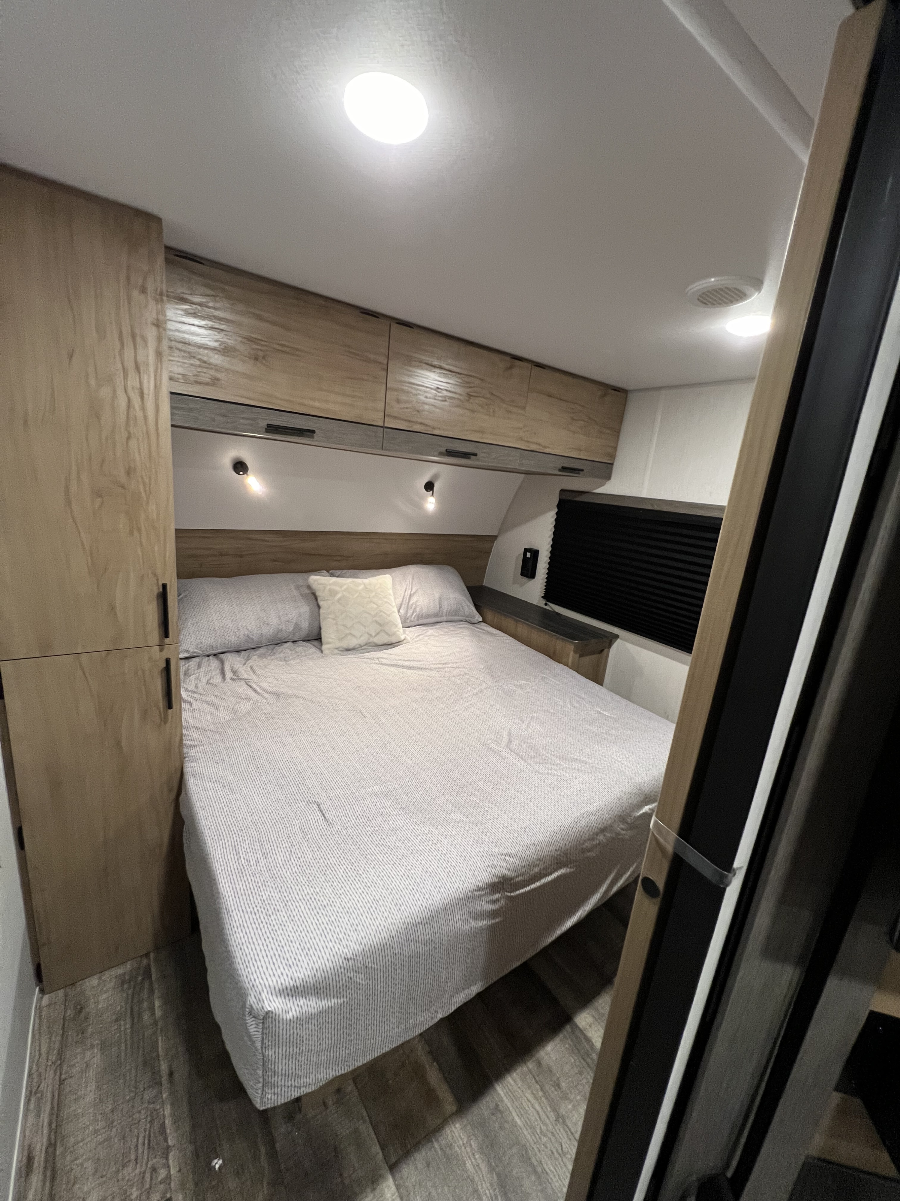 Inside an RV rental: a compact bedroom with a bed, overhead wooden cabinets, modern lighting, and a small bedside area.. Forest River Campsite Reserve 26CJ 2024
