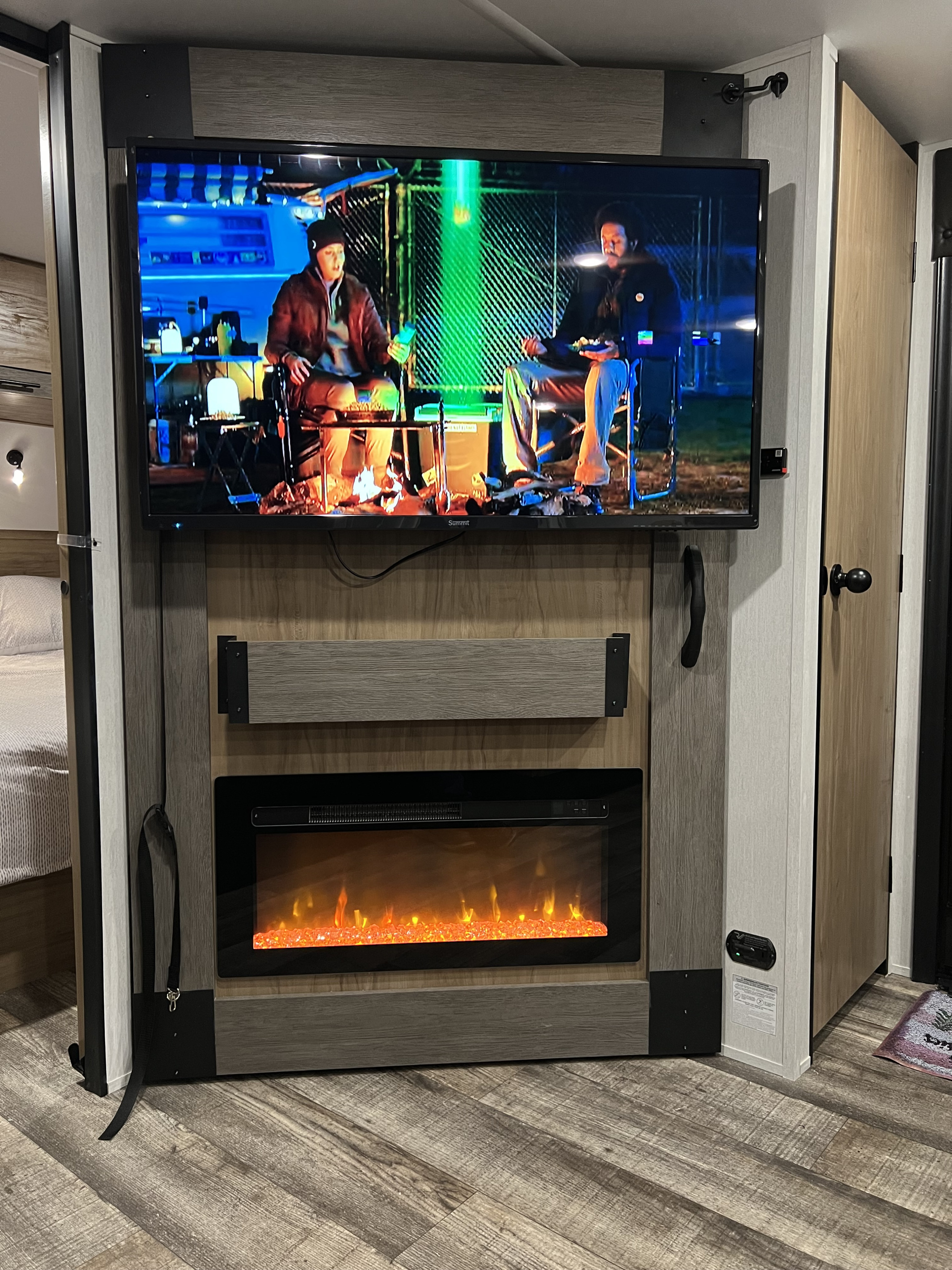 RV rental interior: A mounted TV playing a campfire scene above a glowing electric fireplace, with a bed visible to the left.. Forest River Campsite Reserve 26CJ 2024