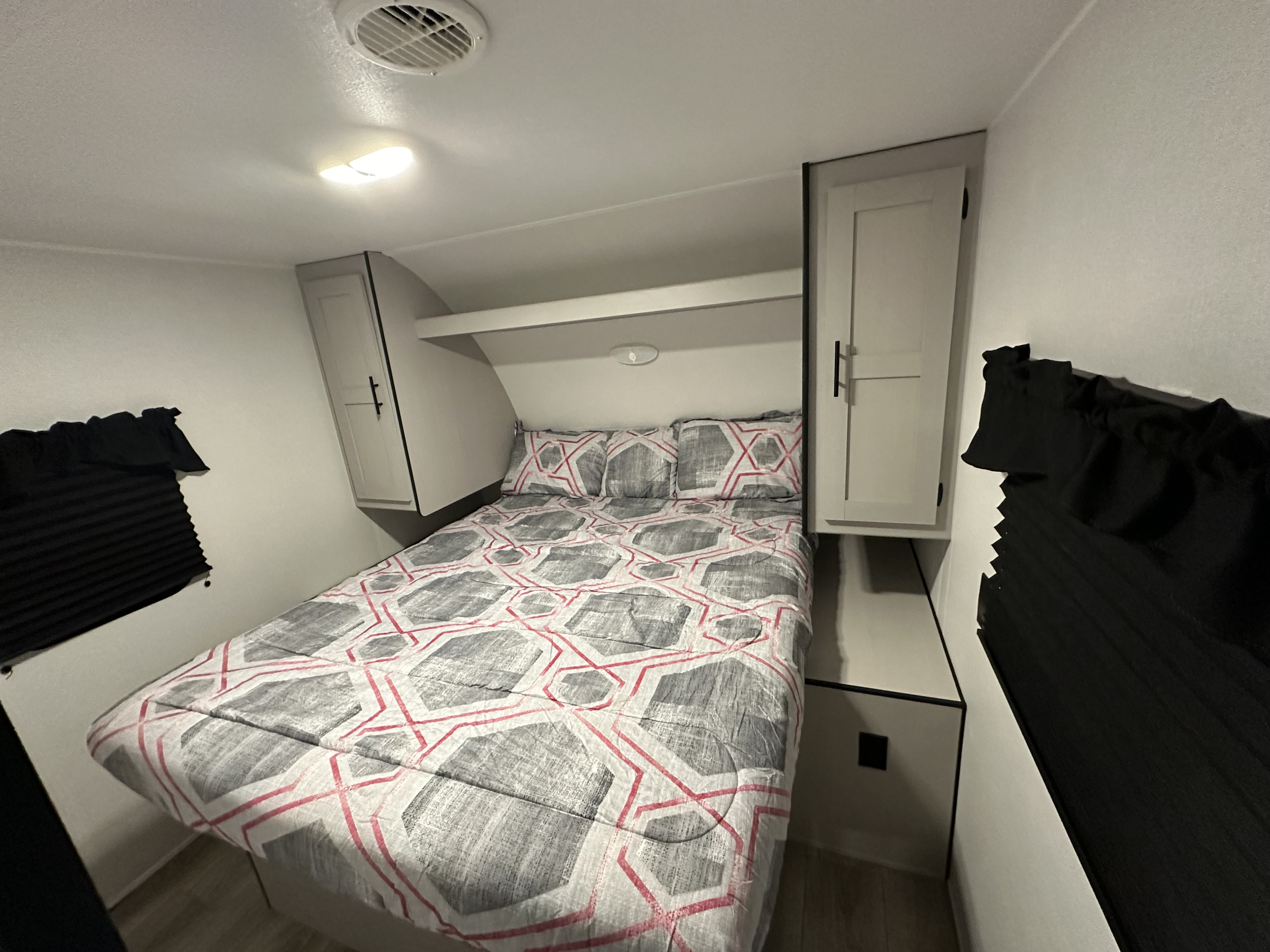 Cozy RV rental bedroom with a grey/red geometric comforter, overhead storage, side cabinets, and window blinds.. Heartland RVs Prowler Lynx 2024
