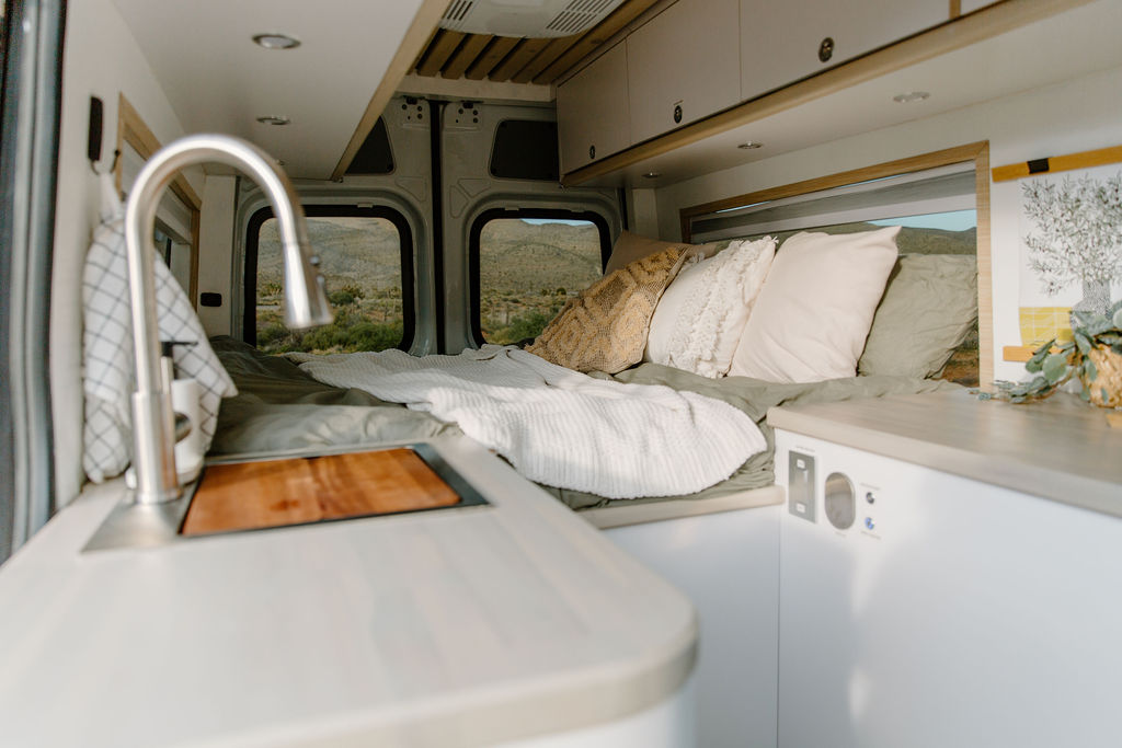 Modern RV rental interior with a cozy bed, equipped kitchen sink, & panoramic desert views through the windows.. Mercedes Sprinter 2021