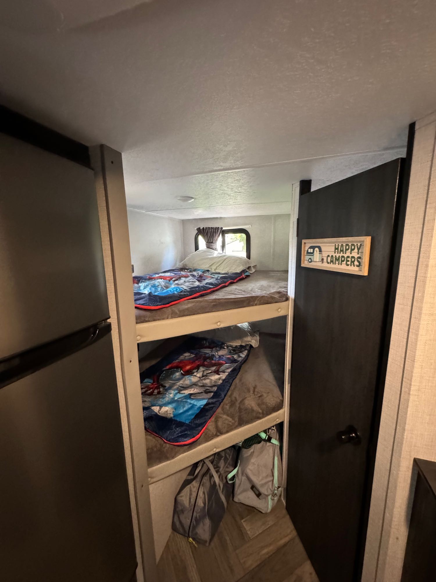 Interior view of an RV rental showing bunk beds with sleeping bags, a refrigerator, and a door with a 'Happy Campers' sign.. Forest River 1680BSK 2021