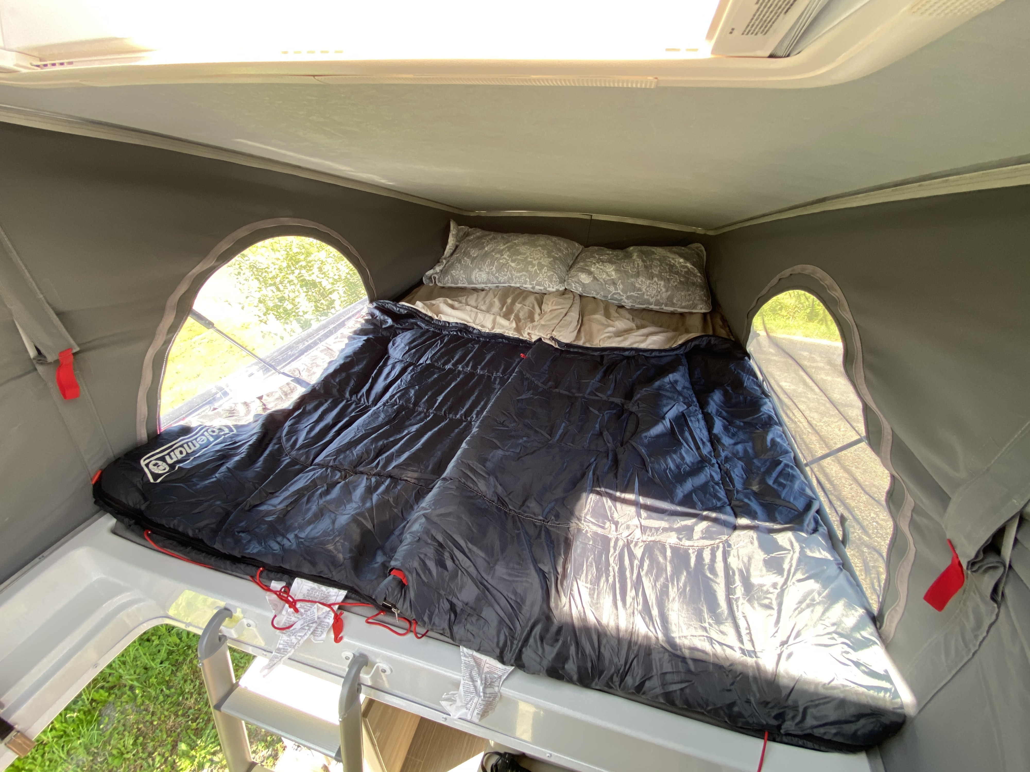Cozy rv rental pop-top bed, showing a Coleman sleeping bag, patterned pillows, and round windows overlooking greenery.. Thor Motor Coach Tellaro 2023
