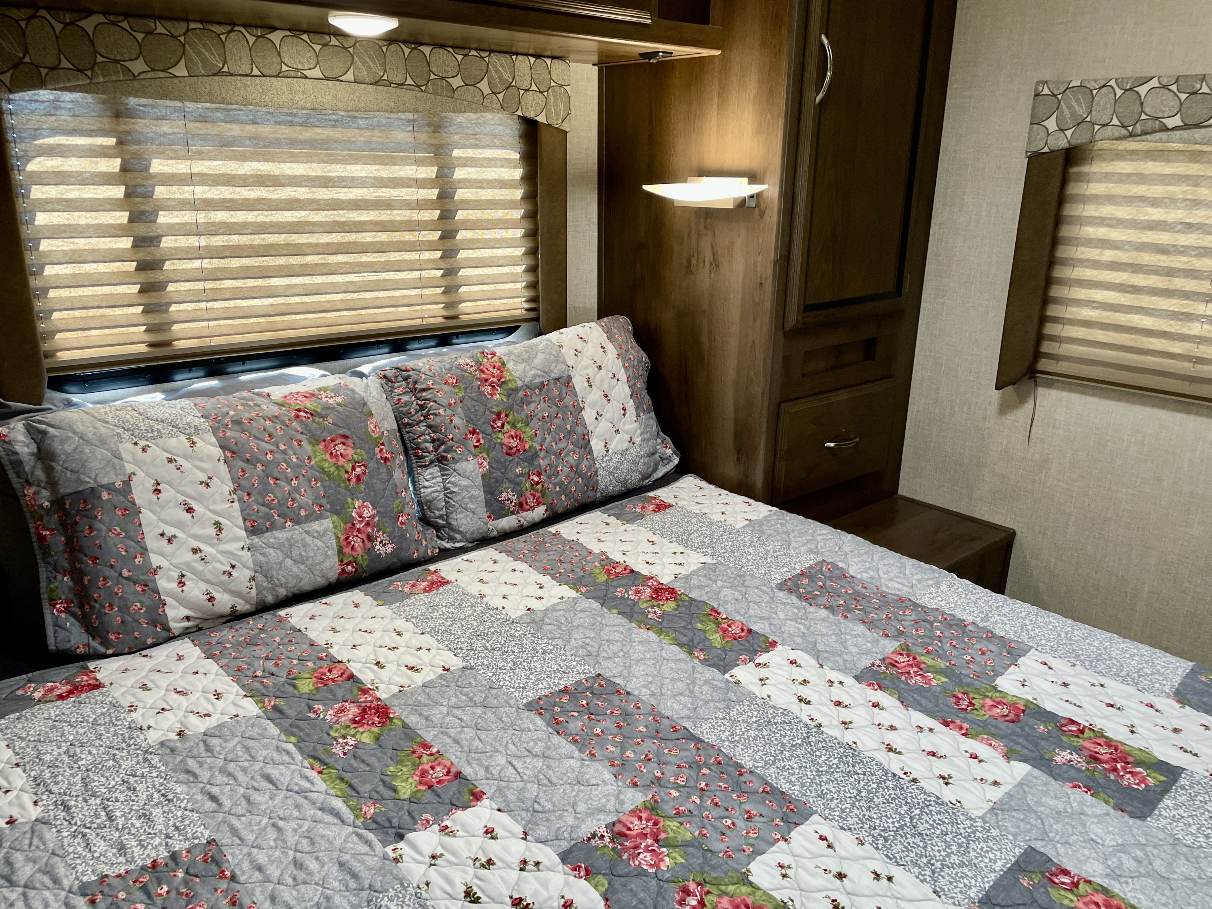 Cozy RV rental bedroom features a floral and grey patchwork quilt, horizontal blinds, and warm wood cabinets.. Coachmen Freelander 2020