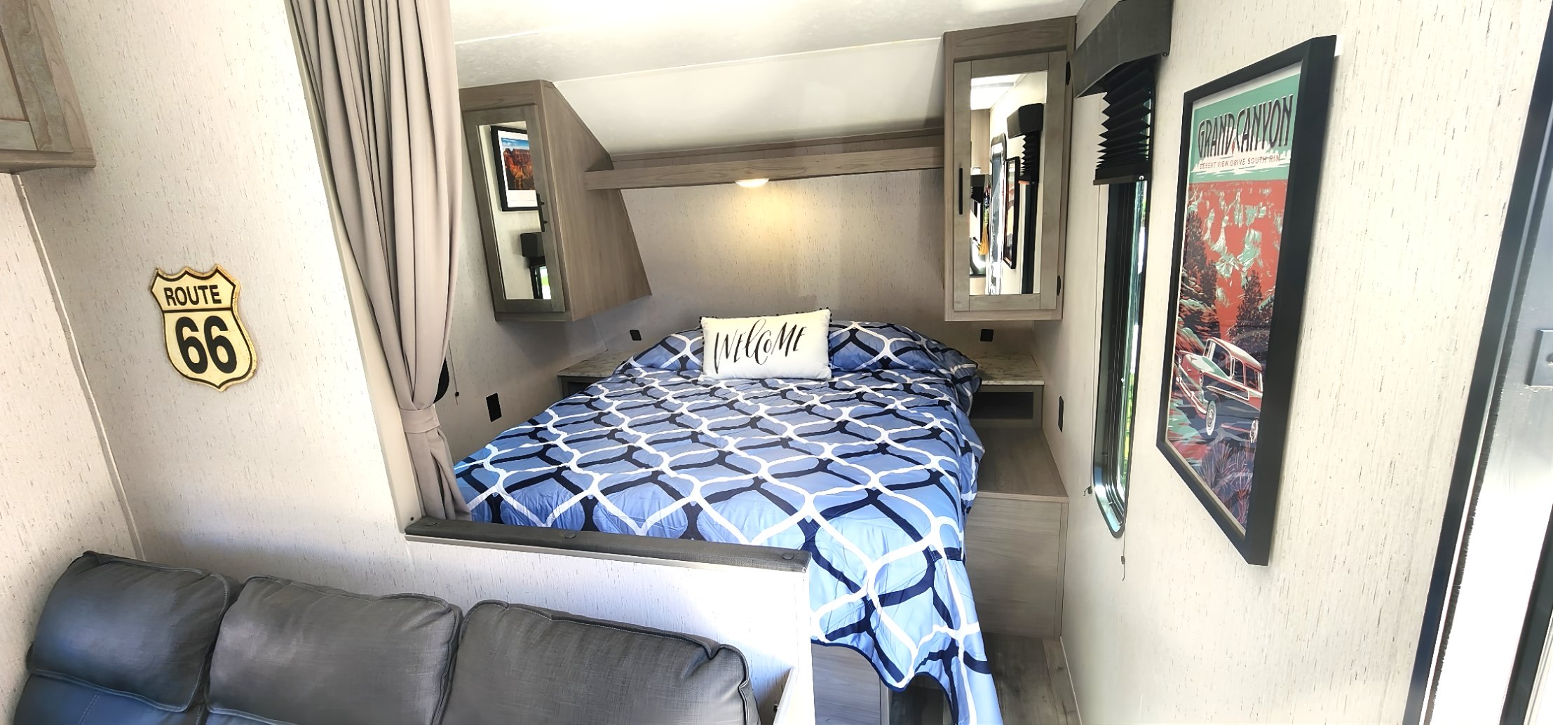 Cozy rv rental bedroom features a blue patterned bed, "Welcome" pillow, a Route 66 sign, and Grand Canyon art.. Dutchmen Coleman Lantern LT 2023