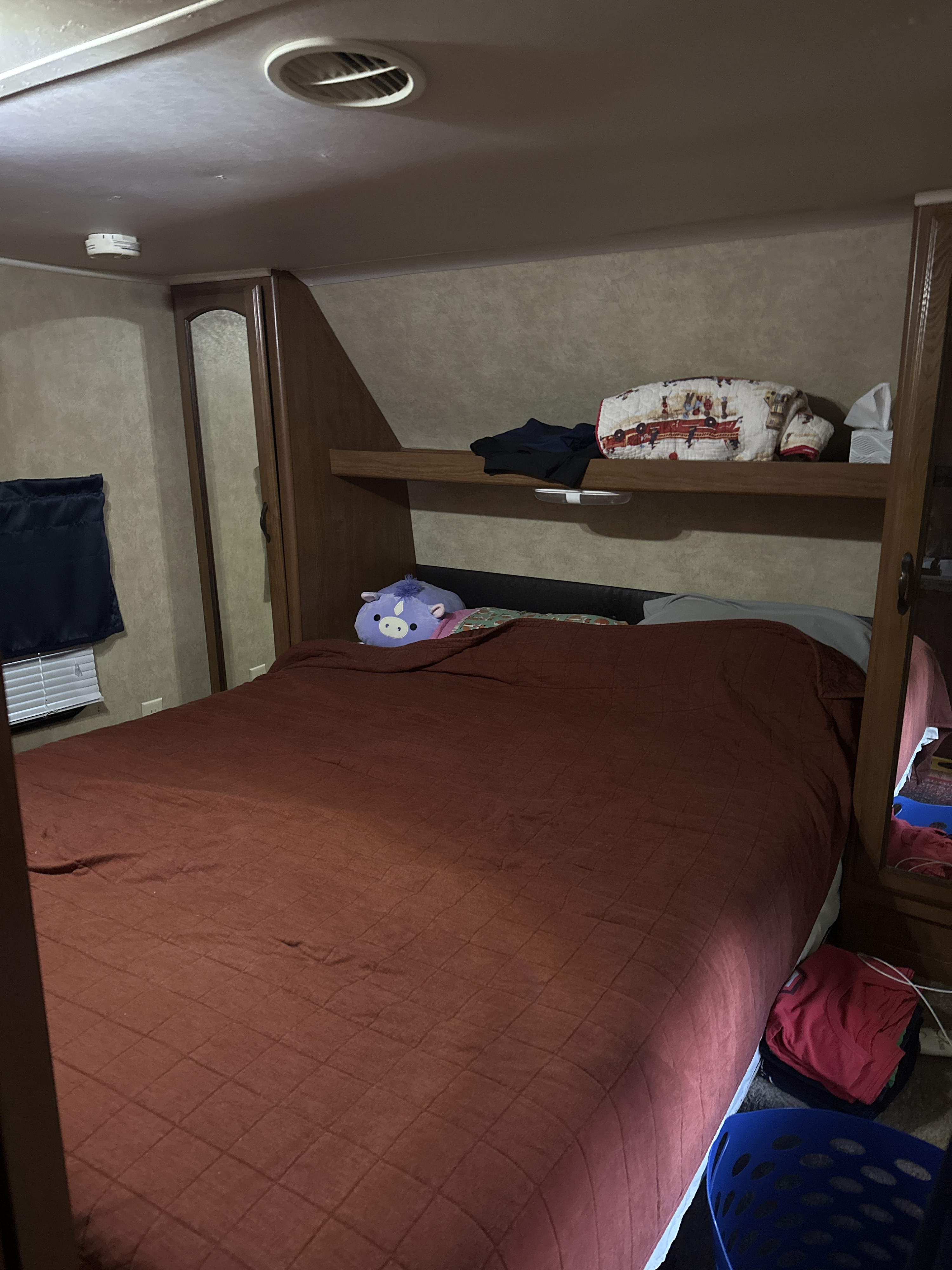 Inside an RV rental bedroom: a cozy bed with red comforter, purple plushie, built-in storage, and window blinds.. Forest River Wildwood 2012