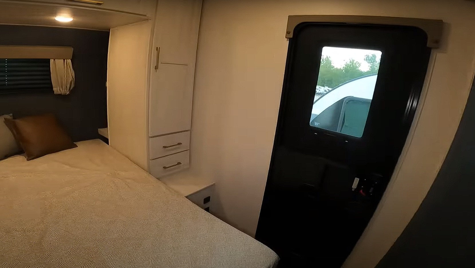 Inside an RV rental bedroom: bed with pillows, built-in wardrobe, and an entry door revealing an outdoor RV park scene.. Forest River Surveyor 296QBLE 2024