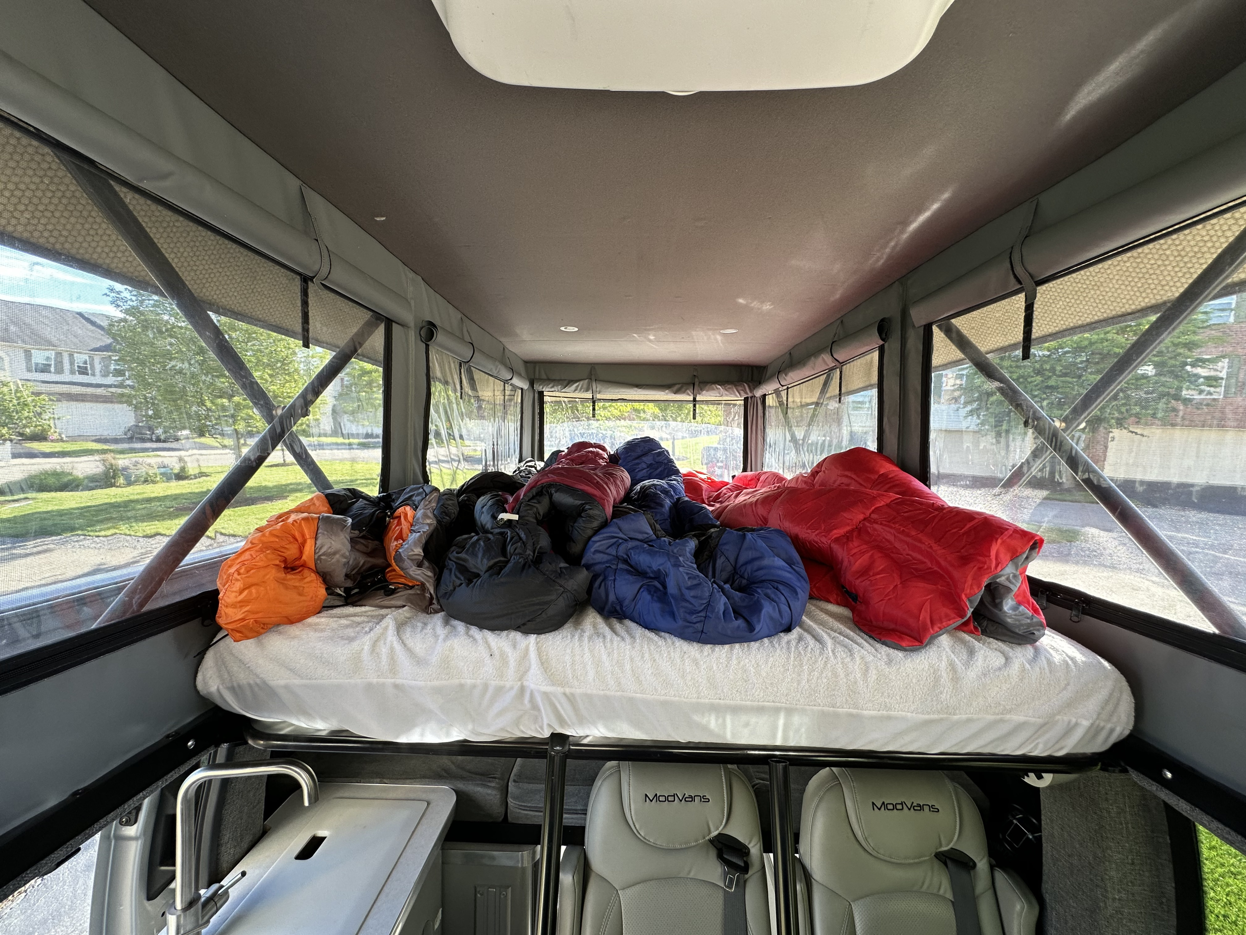Pop-top camper RV rental interior, showcasing a bed with sleeping bags, large windows, and ModVans seats below.. Modvan CV1 Ford Transit 2021