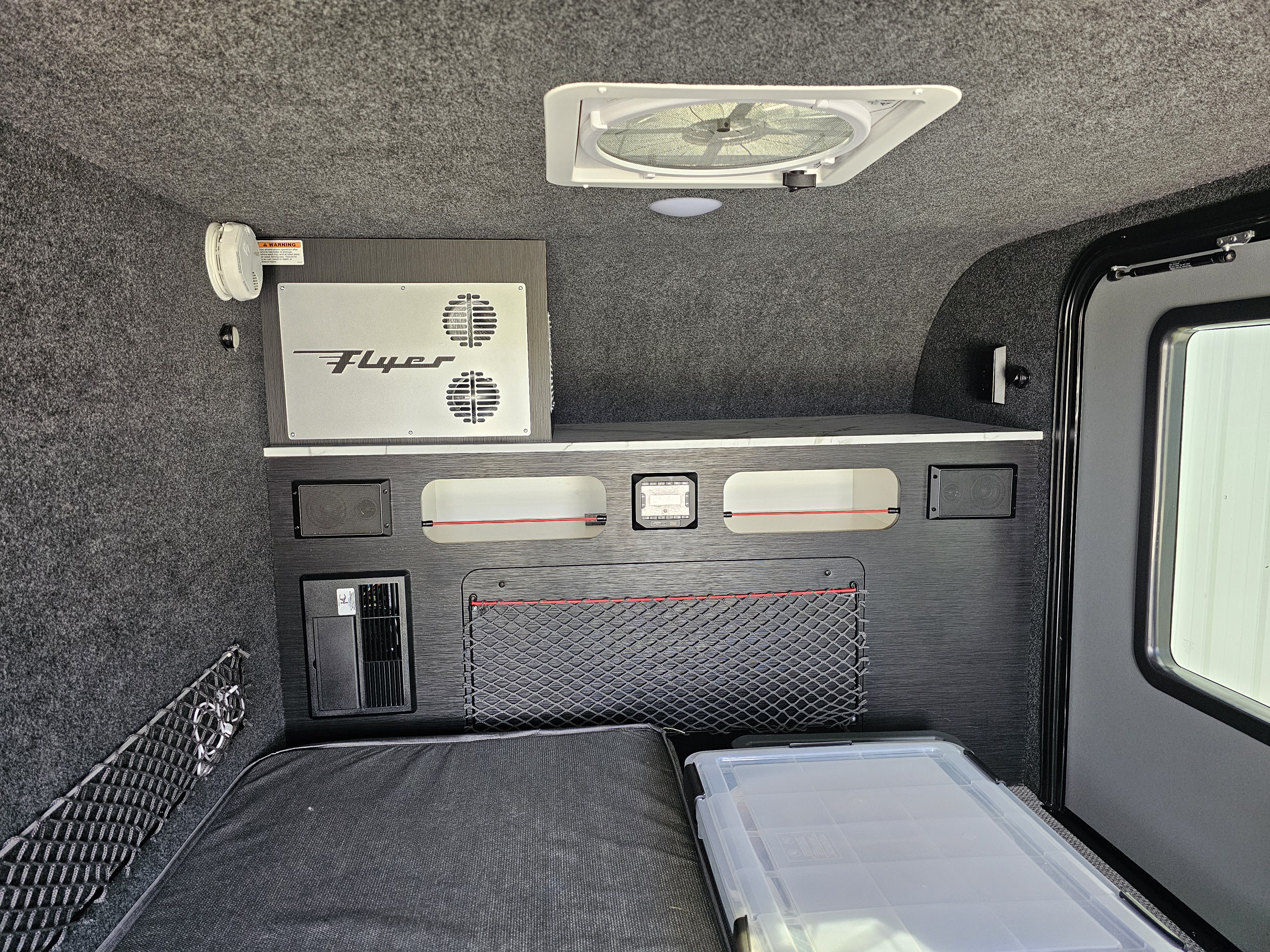 Inside an rv rental, a dark grey interior features a sleeping area, 'Flyer' power unit, ceiling fan, and built-in storage solutions.. InTech RV Flyer 2024