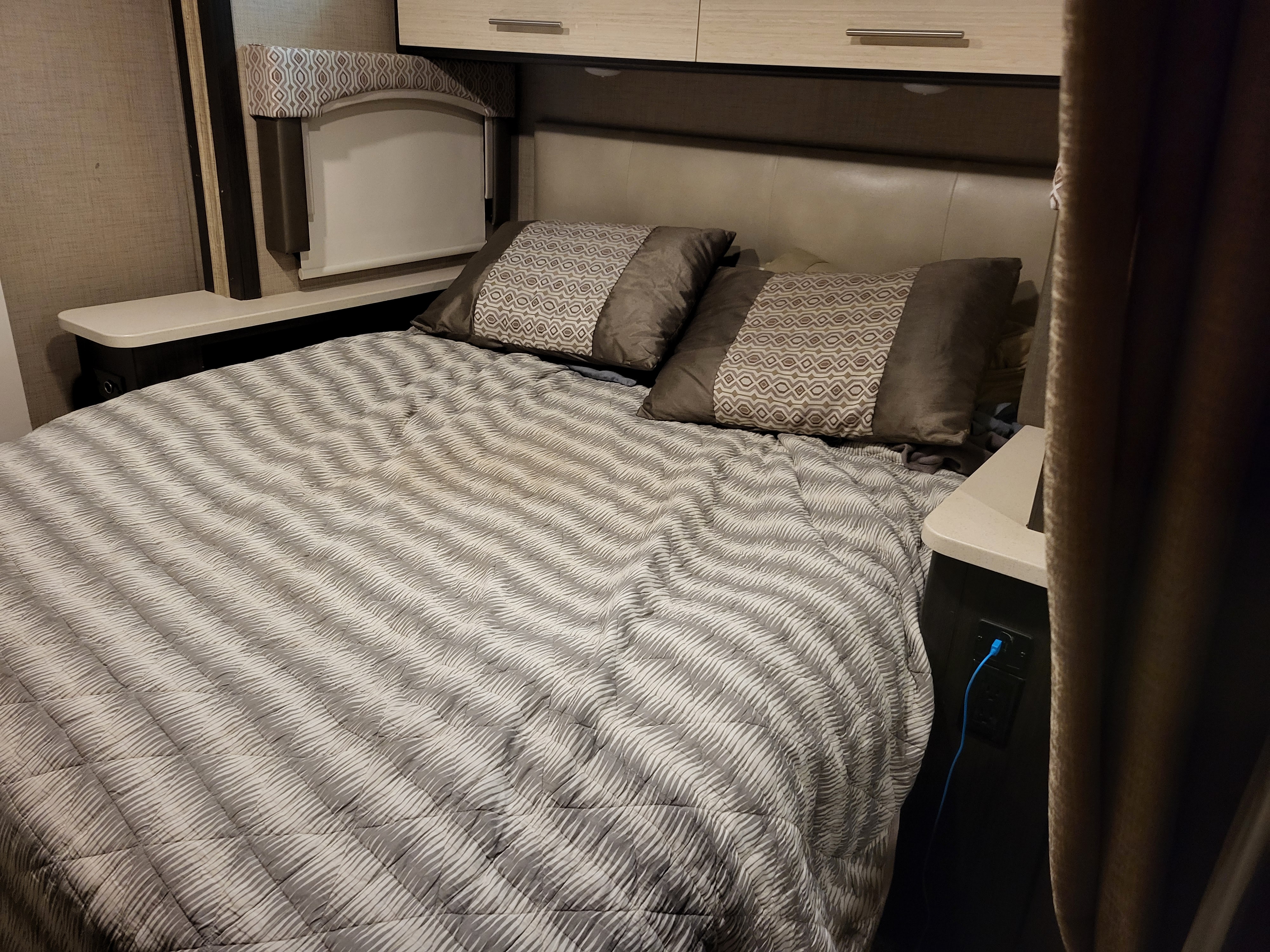 Comfortable RV rental bedroom featuring a striped bed, decorative pillows, overhead cabinets, a window, and power outlet.. Thor Vegas 2020