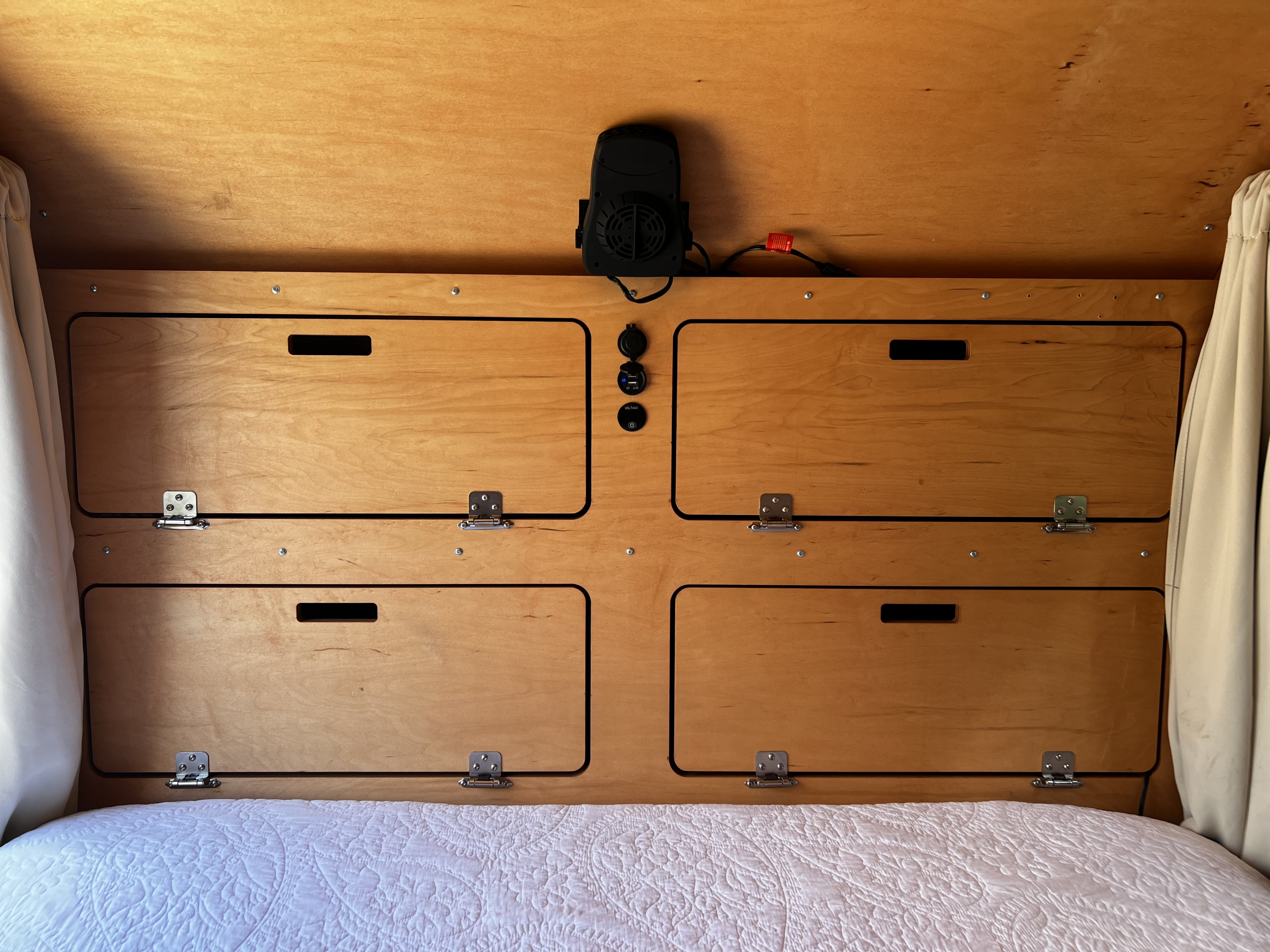 Cozy wooden interior of an RV rental, showing built-in storage, fan, & charging ports above a bed.. TC Teardrop 5x10 Overland 2021