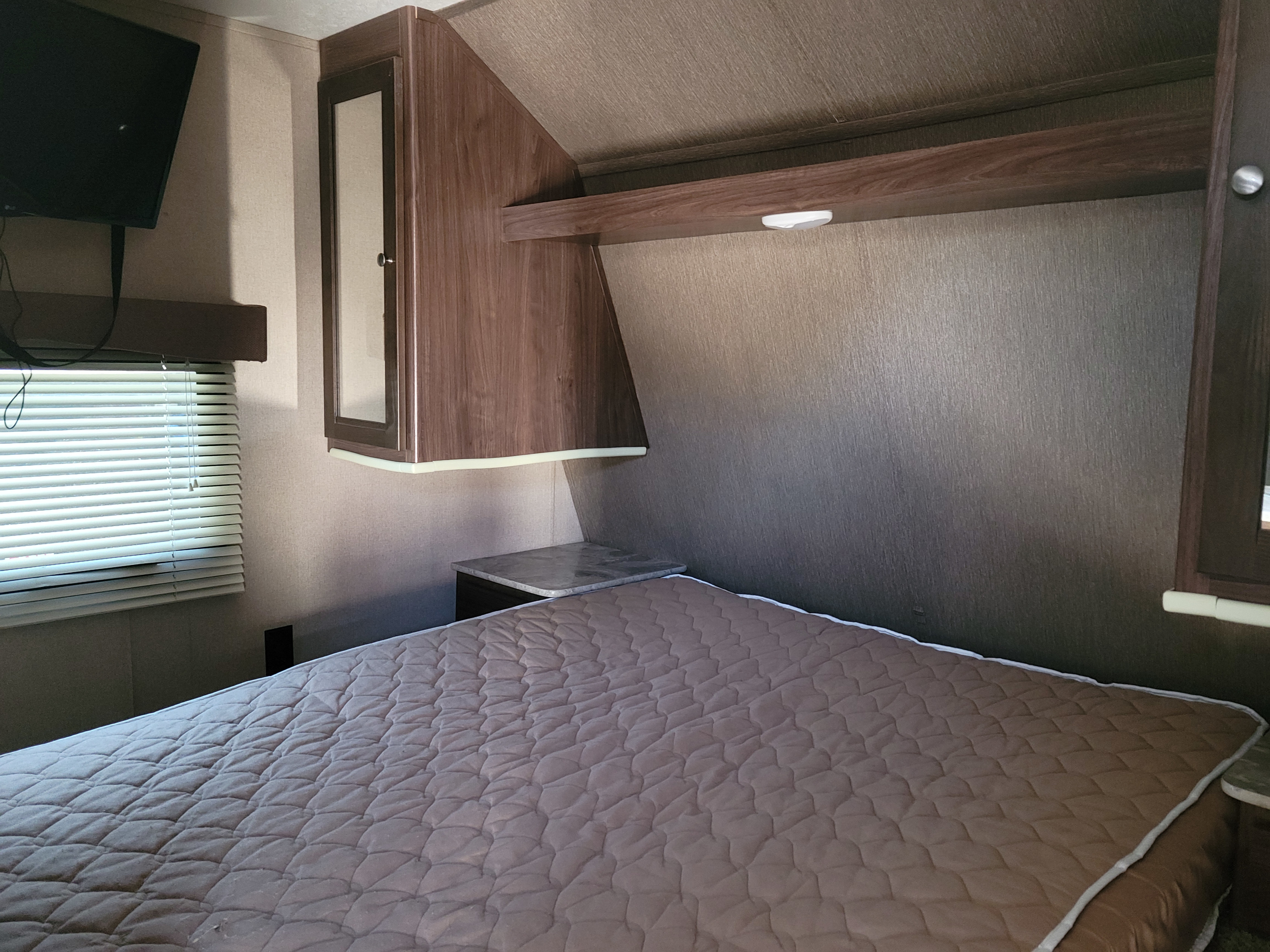 Interior of an RV rental bedroom with a quilted bed, overhead cabinets, wall-mounted TV, and window blinds.. Coleman Lantern 2018