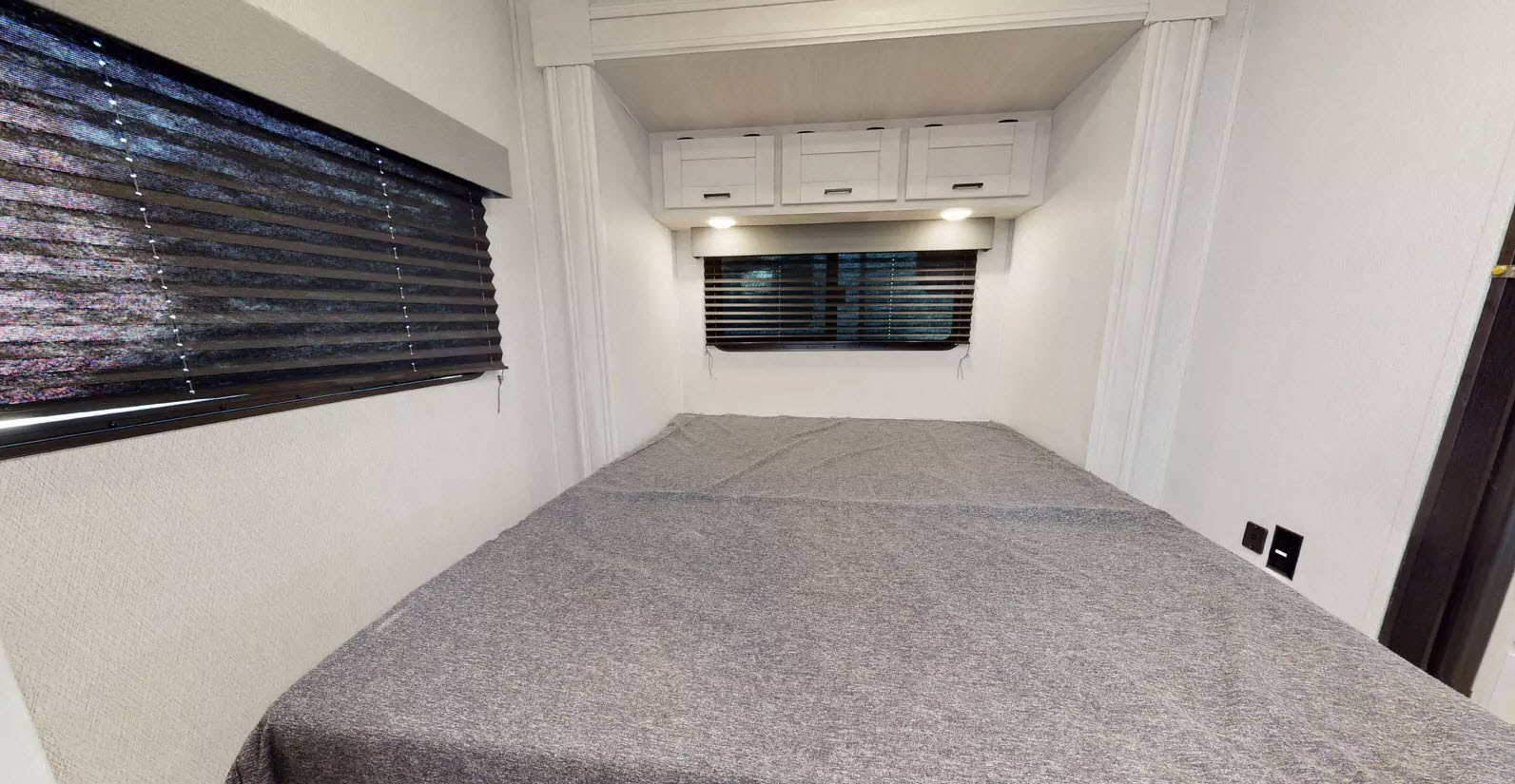 RV rental bedroom features a gray bed, window blinds, and overhead cabinets, offering a cozy, compact space.. East to West 220SC 2025