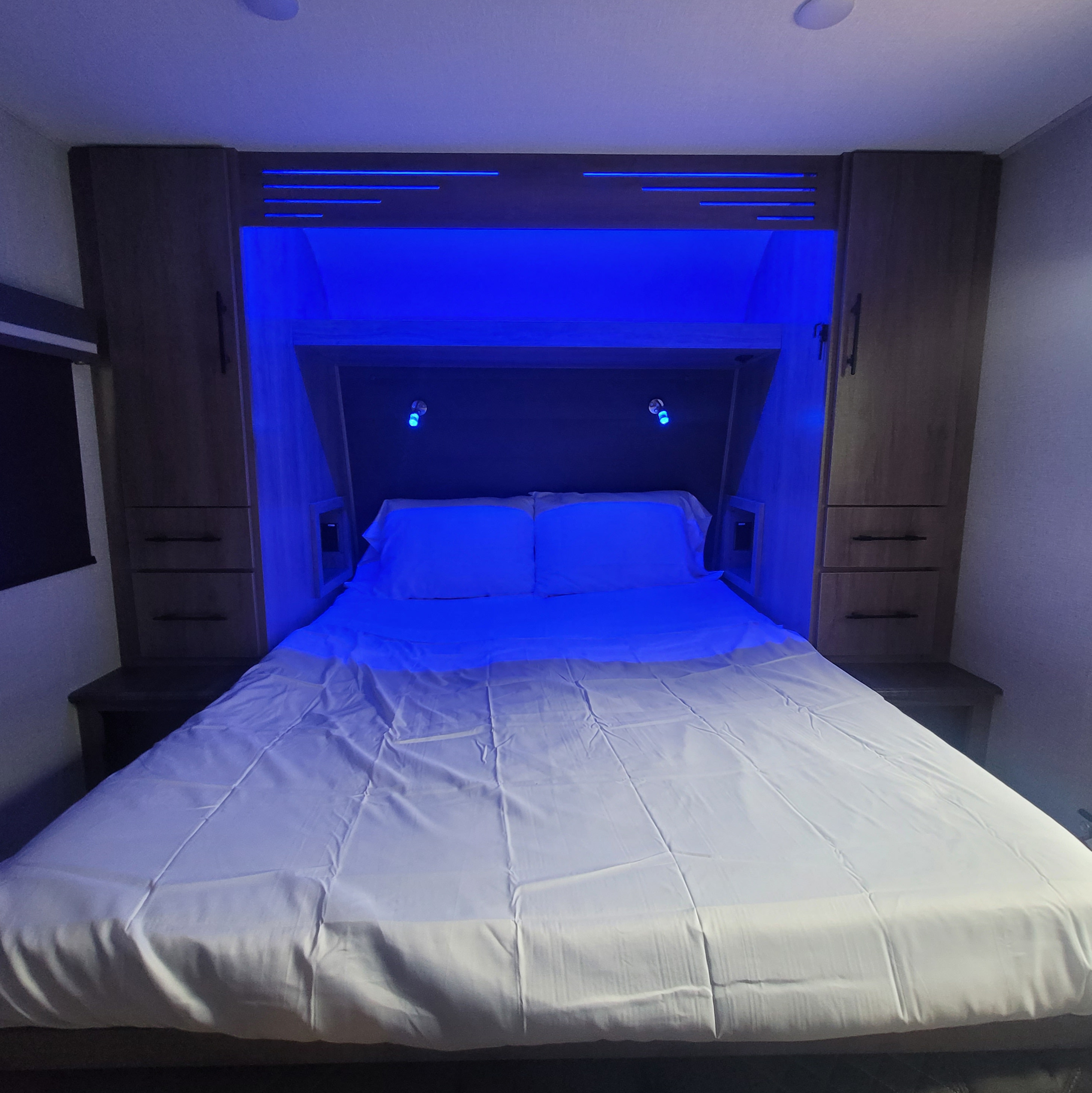 RV rental bedroom with vibrant blue LED lighting, a queen bed, built-in side cabinets, and reading lamps.. Grand Design Imagine AIM 2023