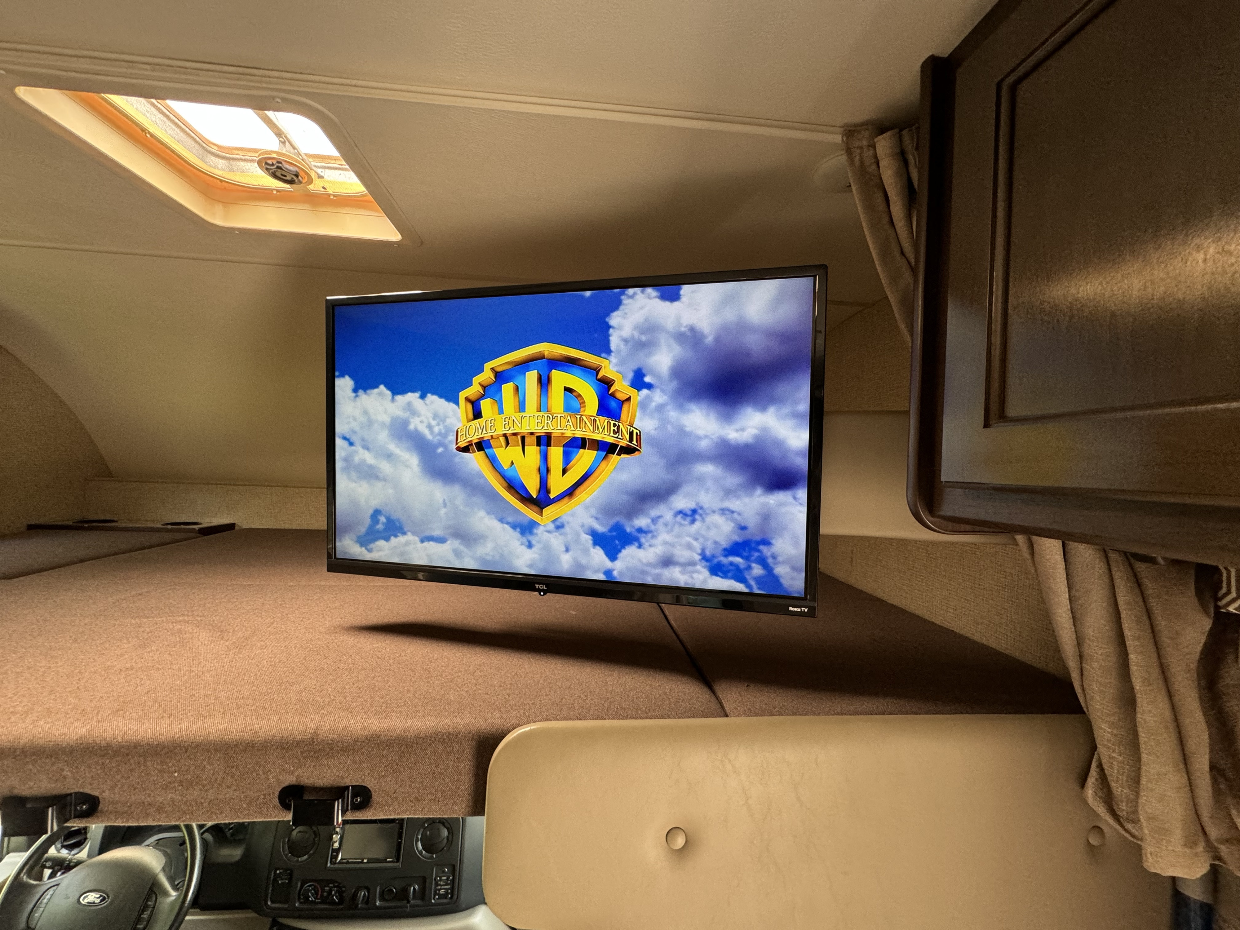 Cozy RV rental interior with a TV displaying Warner Bros. Home Entertainment, a skylight, and a bed above the vehicle's cockpit.. Thor Four Winds 2018