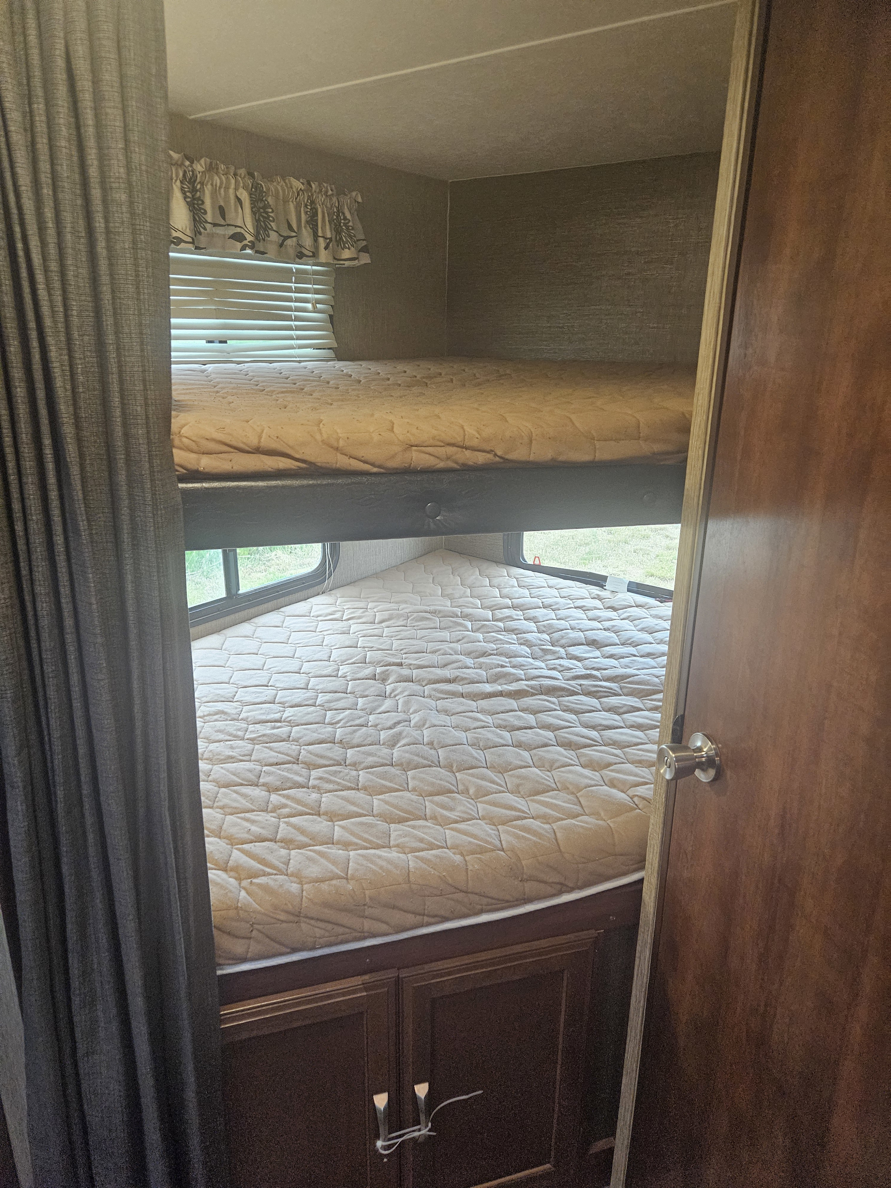 Inside an RV rental: a bunk bed setup with two mattresses, windows, blinds, and lower cabinets. Dark curtain and door on right.. Keystone RV Passport Grand Touring 2018