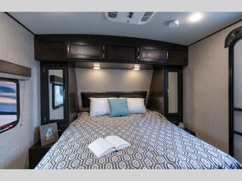 Cozy RV rental bedroom with patterned bed, open book, overhead storage, and scenic window view. Ideal for travel.. Grand Design Imagine 2019
