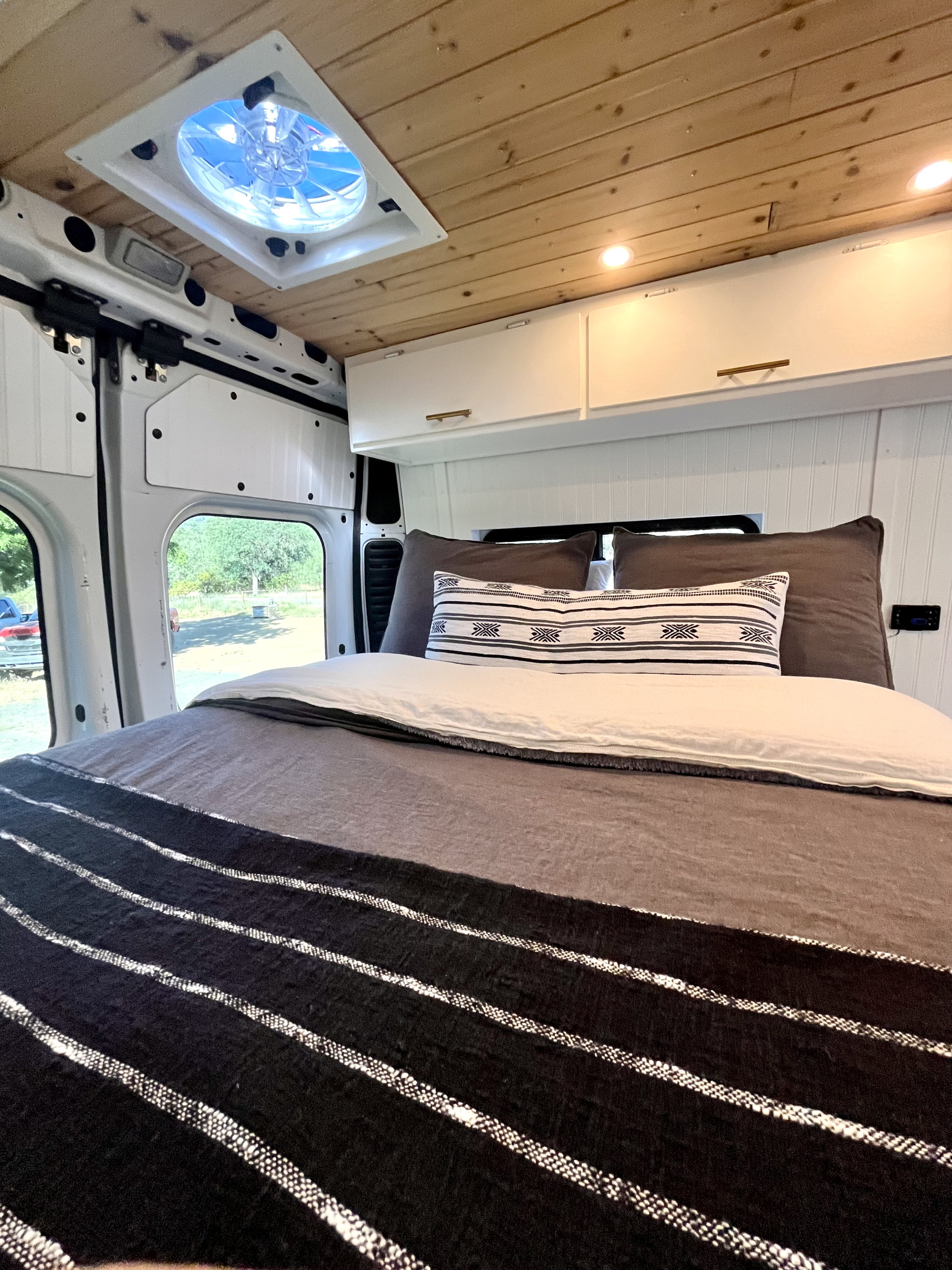 RV rental interior featuring a cozy bed, wooden ceiling with fan, and overhead cabinets for storage.. Dodge B Van 2015