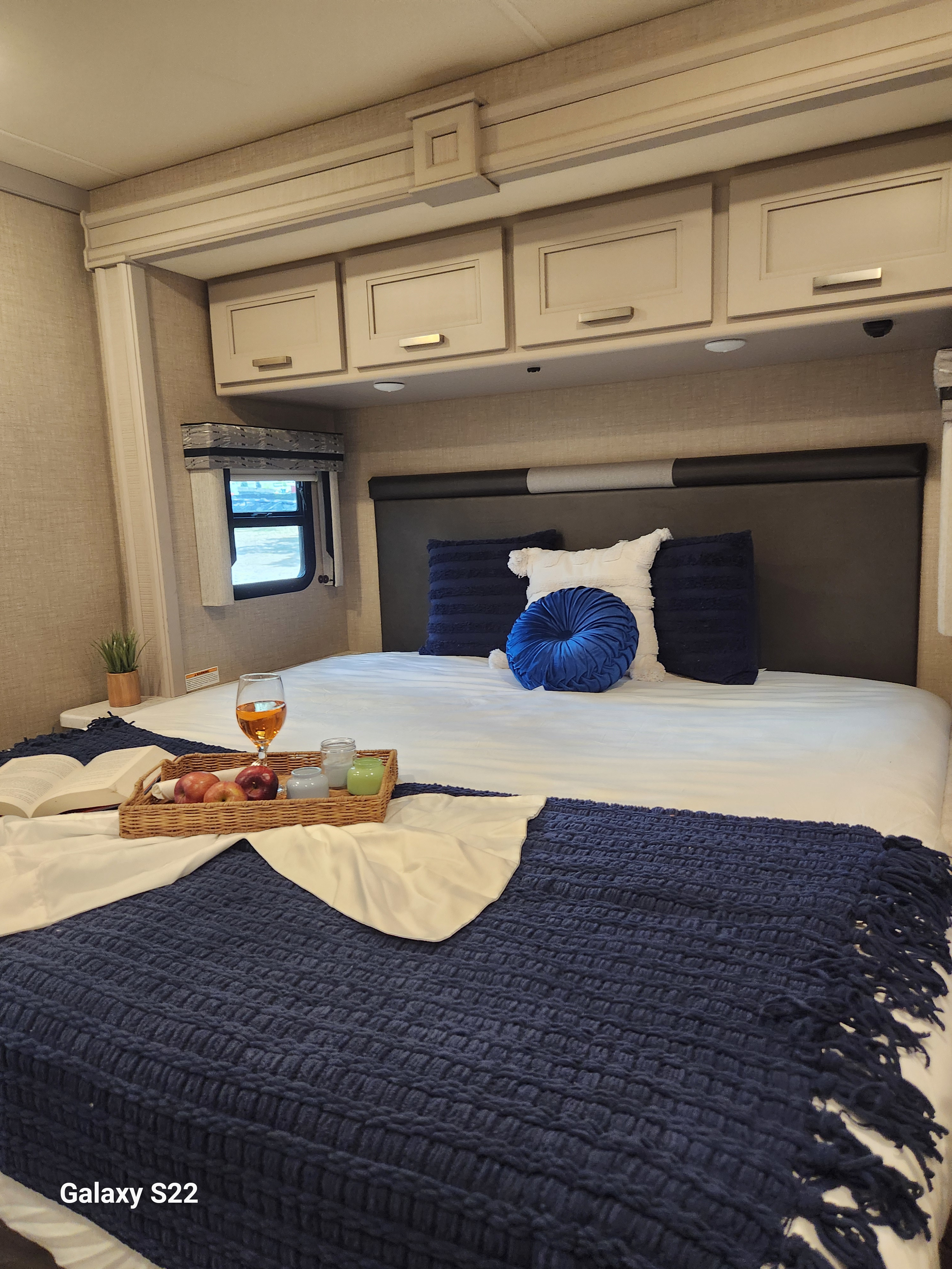 Cozy RV rental bedroom features a bed with blue and white bedding, overhead cabinets, a small window, and a breakfast tray.. Thor Miramar 2023