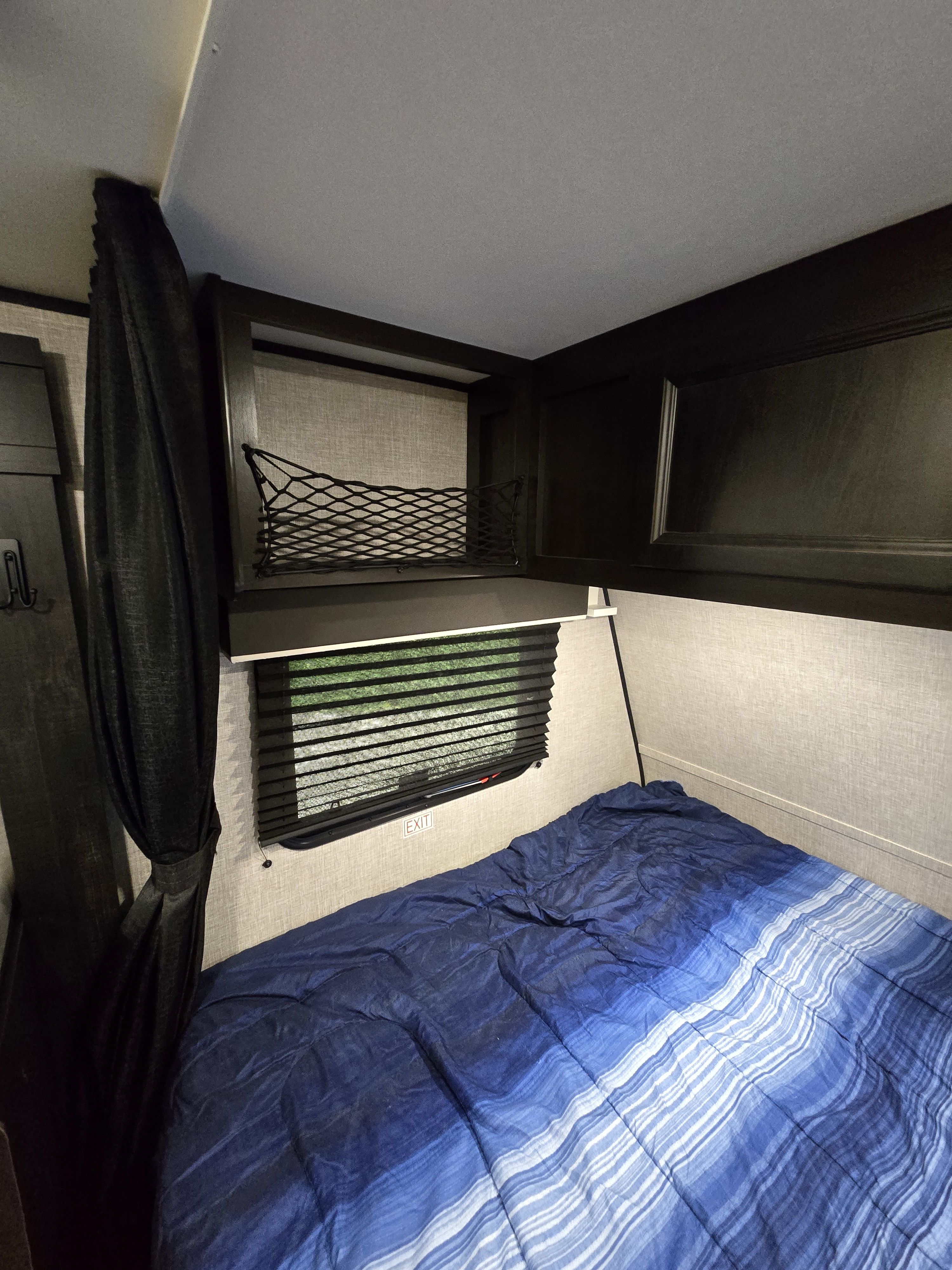 Compact RV rental bedroom featuring a blue striped bed, window with blinds, overhead cabinets, and a mesh storage net.. Jayco Jay Flight SLX 2022