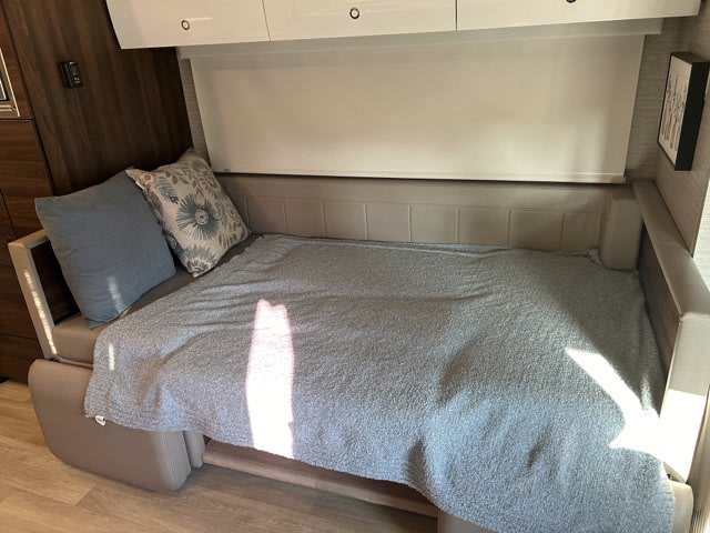 A compact RV rental living space, showing a beige pull-out sofa-bed covered with a soft blue blanket and pillows, illuminated by sunlight.. Winnebago View 2023