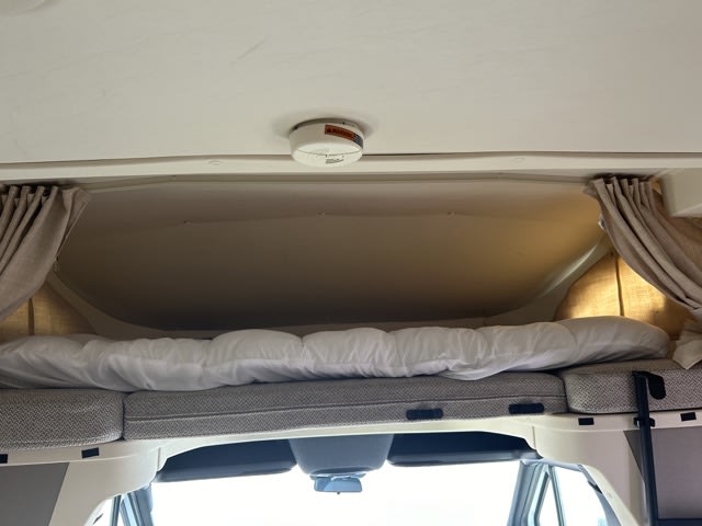 Overhead bunk in an RV rental: a comfy sleeping area with white comforter, side curtains, and a ceiling smoke detector.. Winnebago View 2023