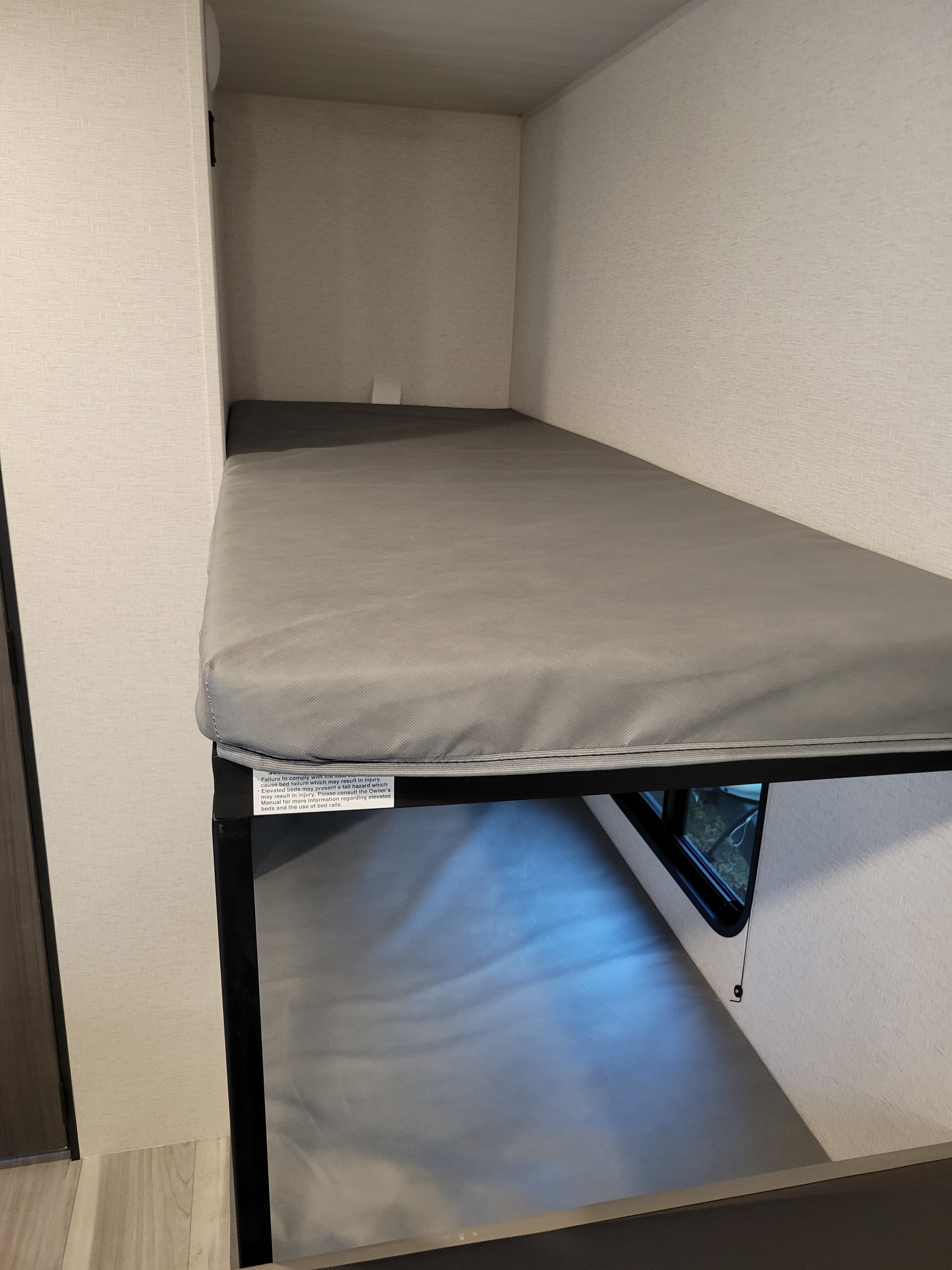 Compact RV rental bunk bed with a gray mattress and safety warning label, designed for efficient space use.. Keystone RV Hideout Sport 2024