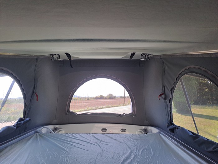 Interior of an RV rental pop-top camper, showing the bed and panoramic windows with an outdoor view of a road and fields.. Bürstner Campeo 600 2024