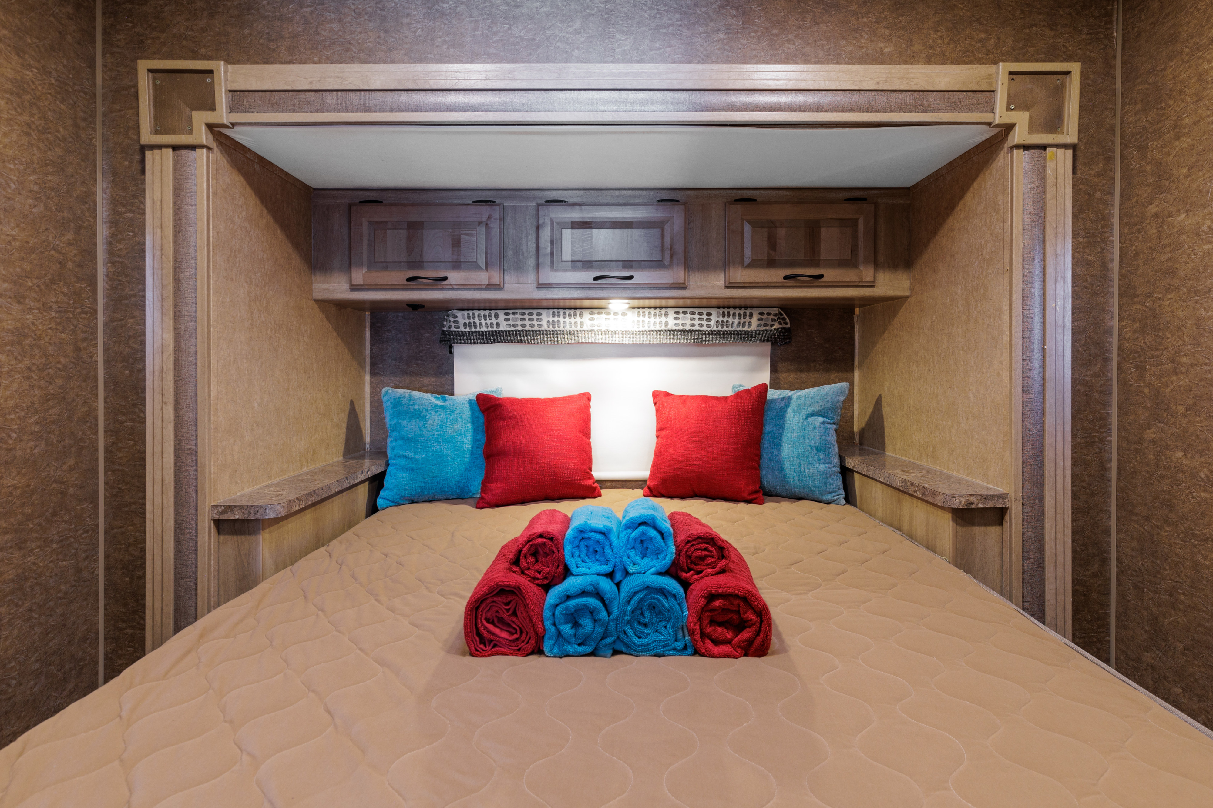 Experience comfort in this RV rental bedroom with a large bed, red & blue pillows, rolled towels, and overhead cabinets.. Coachmen Prism Select 2018