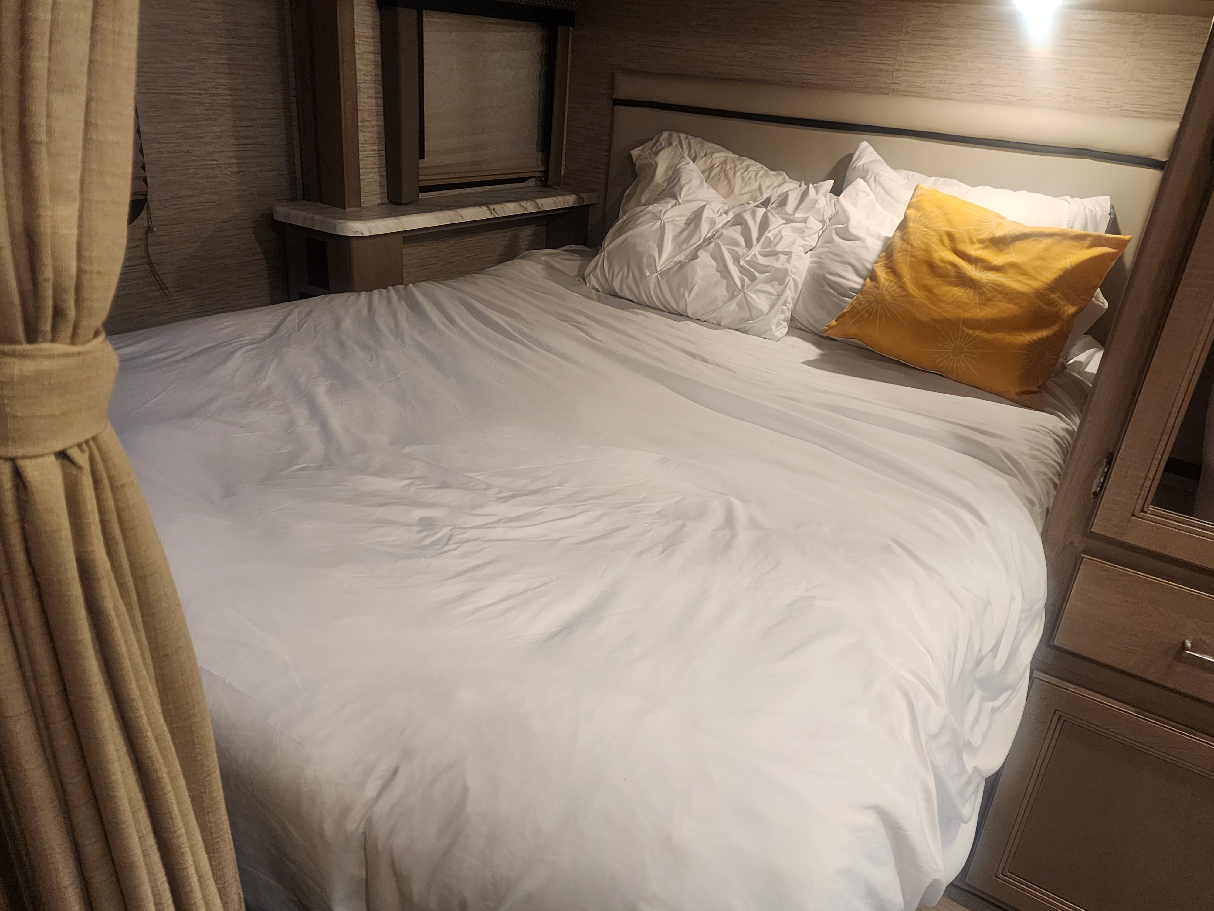 RV rental bedroom: cozy bed with white sheets & yellow pillow, wood cabinetry, and curtains.. Thor Coleman 2022