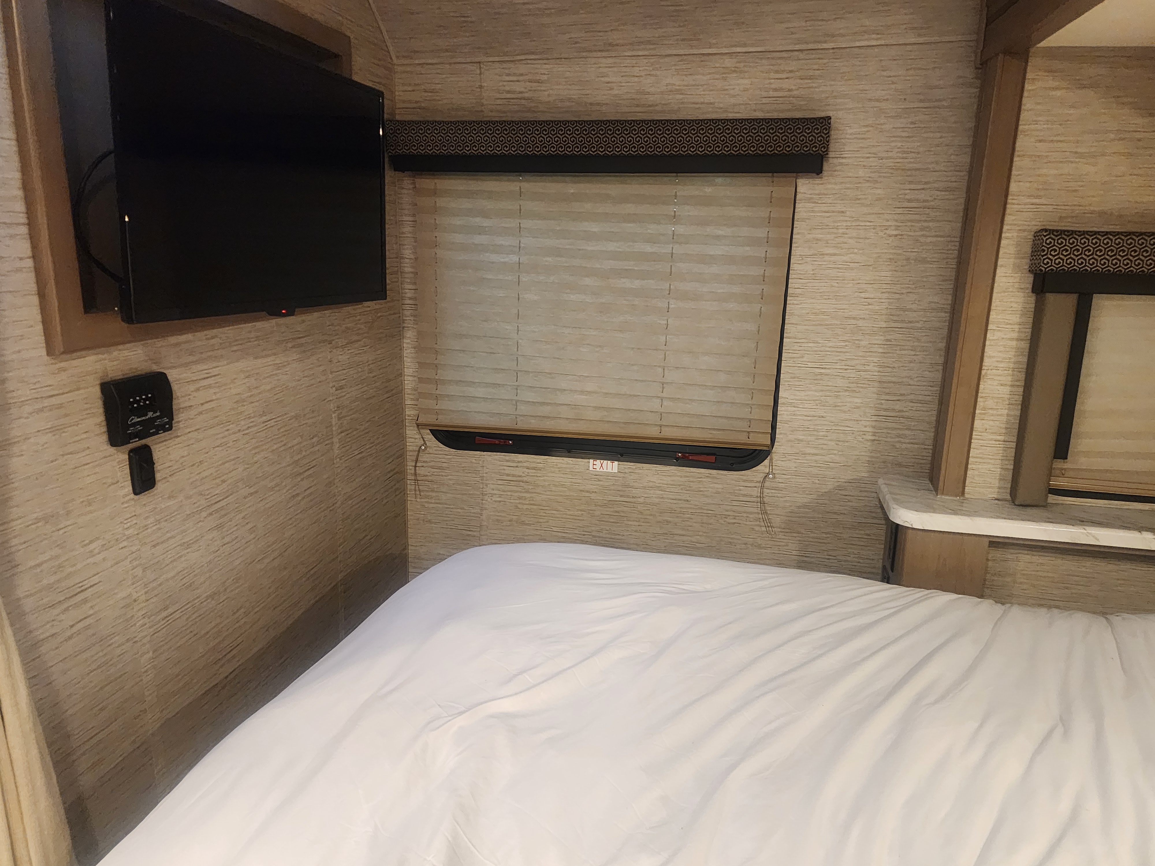 Cozy RV rental interior: A bedroom with a neatly made white bed, wall-mounted TV, and window with blinds & "EXIT" sign.. Thor Coleman 2022