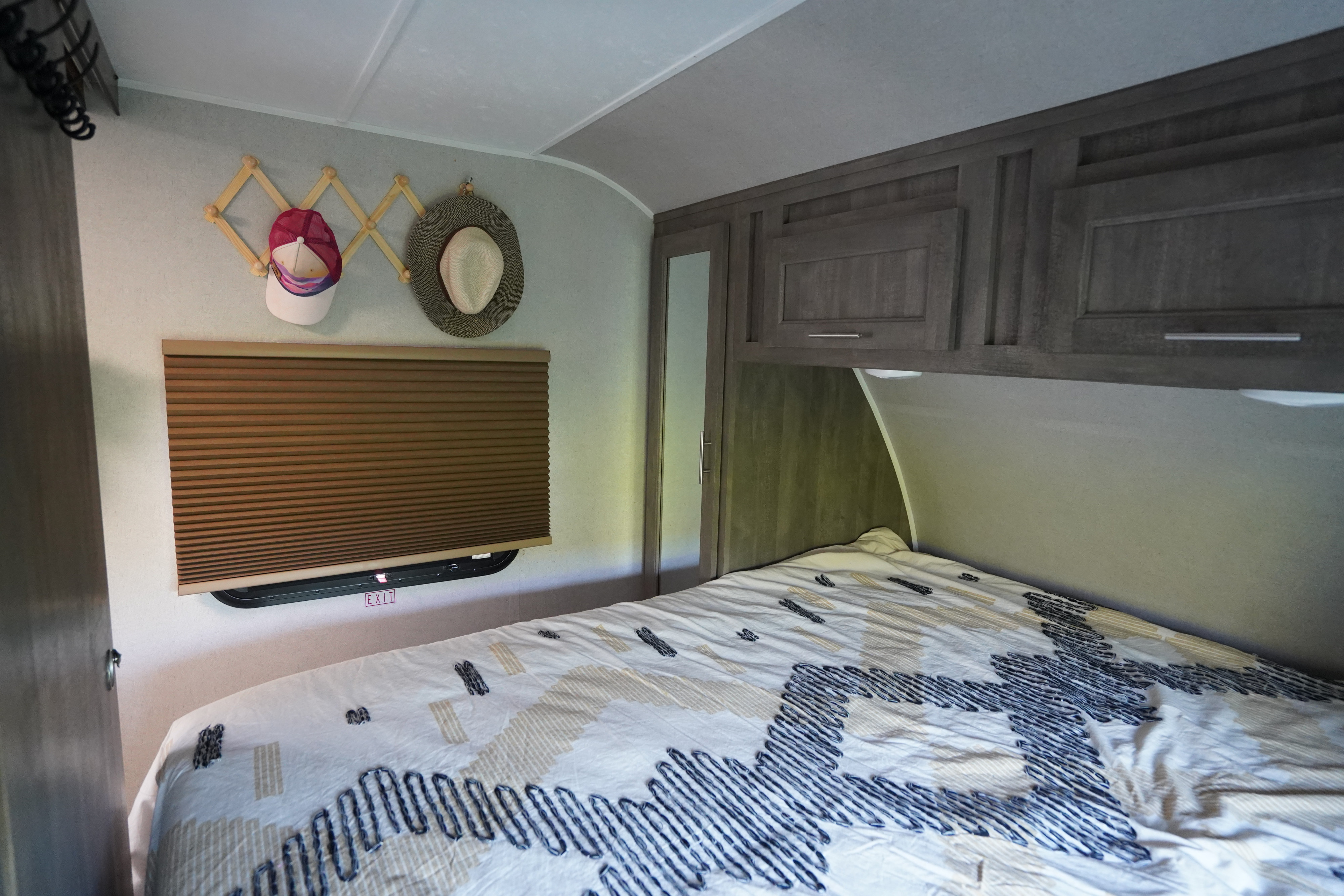 An RV rental bedroom featuring a bed, overhead cabinets, window with blind & exit sign, plus a hat rack.. Forest River Vibe 2020