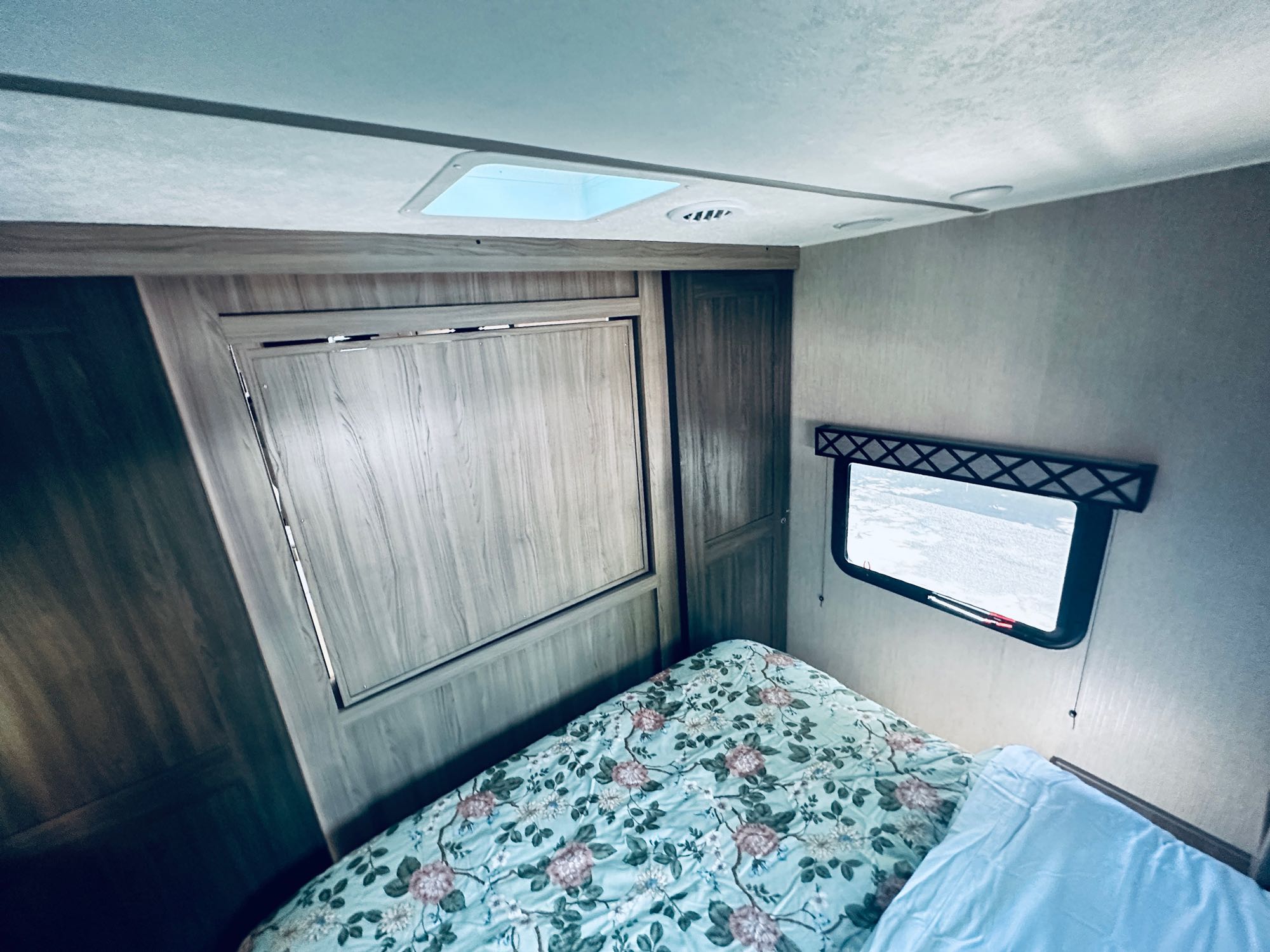 Inside an RV rental bedroom featuring a floral-patterned bed, a window with a snowy view, ceiling skylight, and wood-paneled storage.. Coachmen Freedom Express Ultra Lite 2021