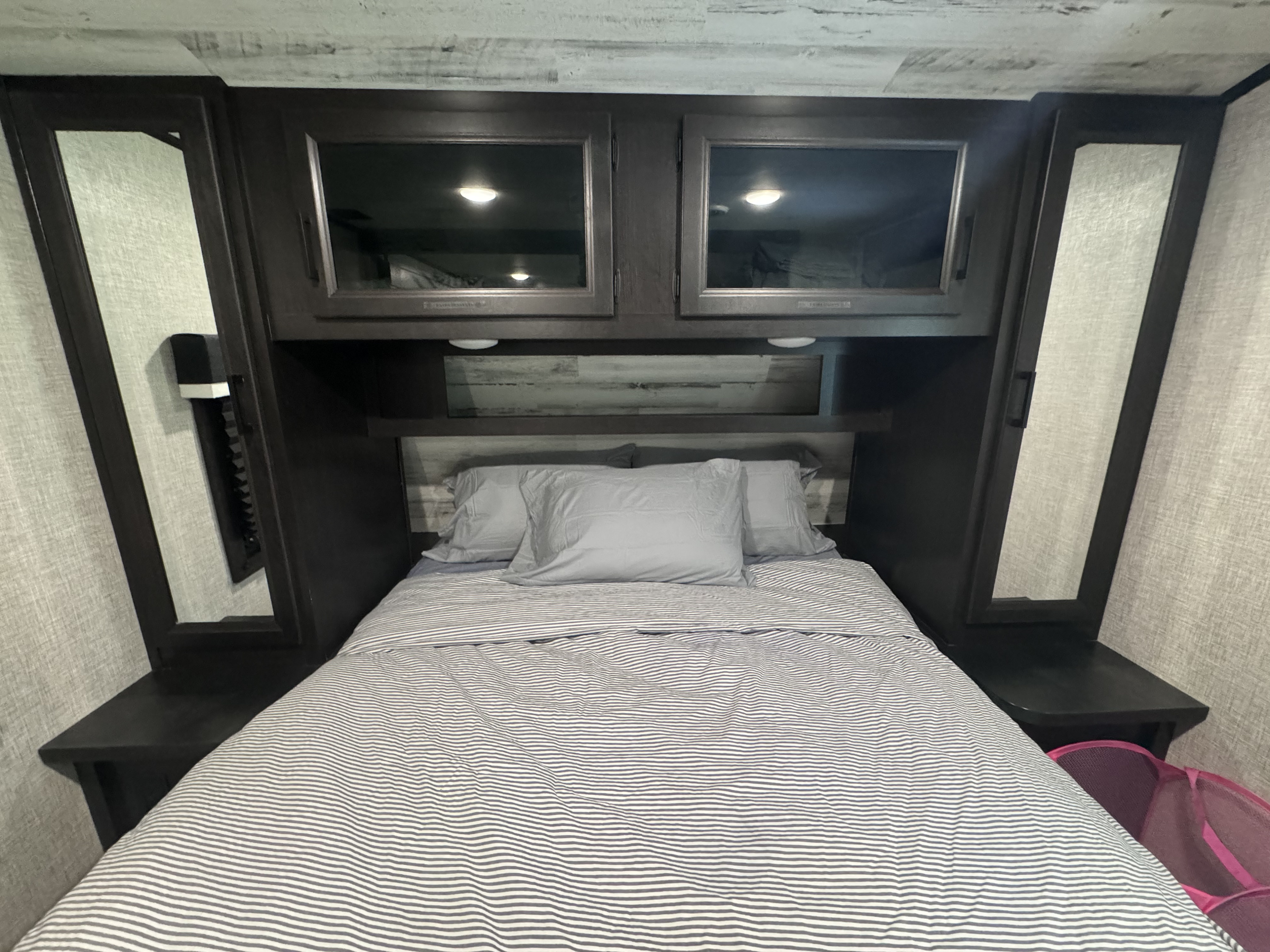 This rv rental features a cozy bedroom with a striped bed, overhead cabinets, and built-in bedside storage.. Jayco Jay Feather 2023