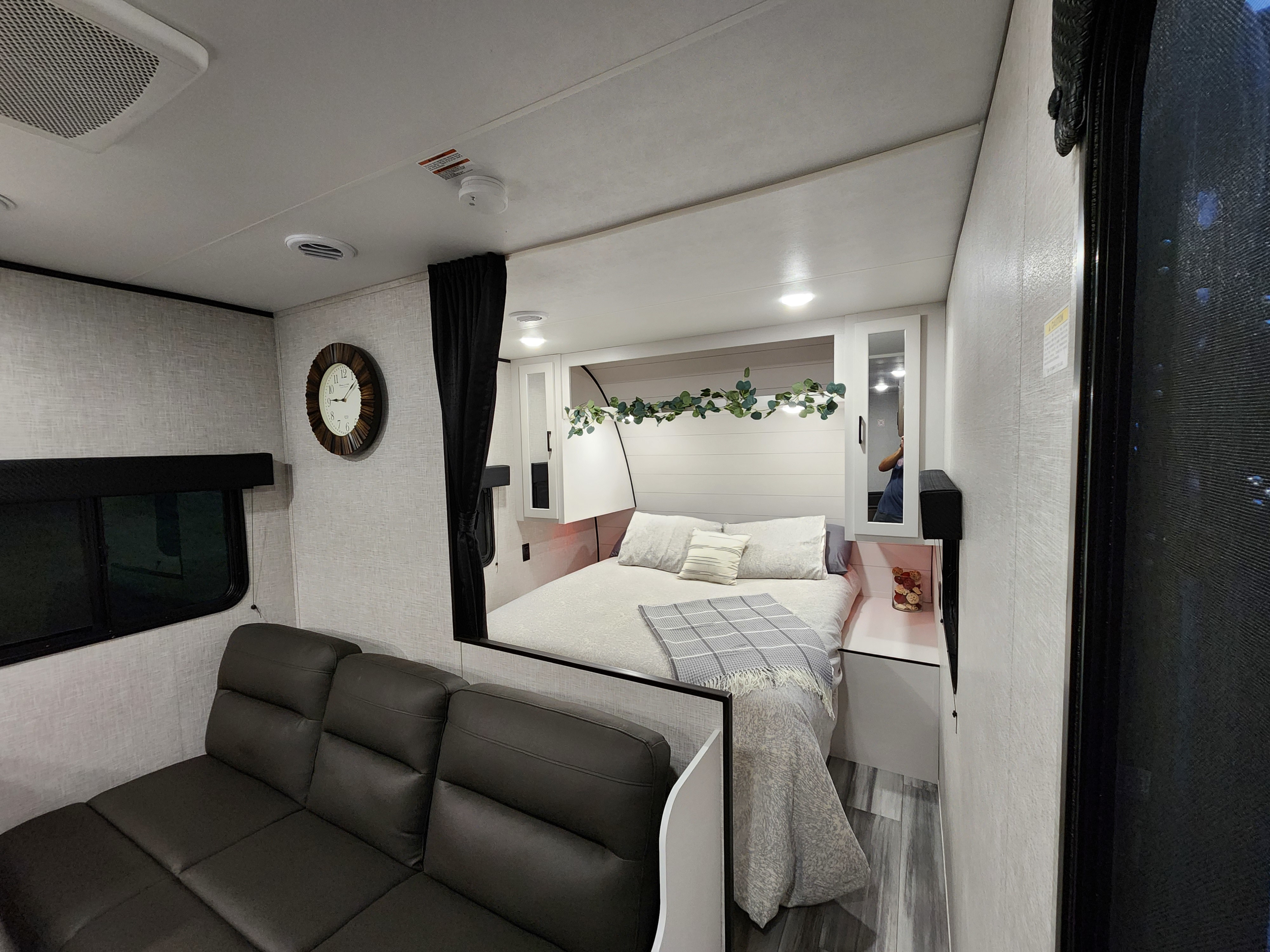 Modern RV rental interior: view of the comfortable bed with shiplap and greenery, plus a dark sofa in the living space.. Jayco Jay Flight 2024