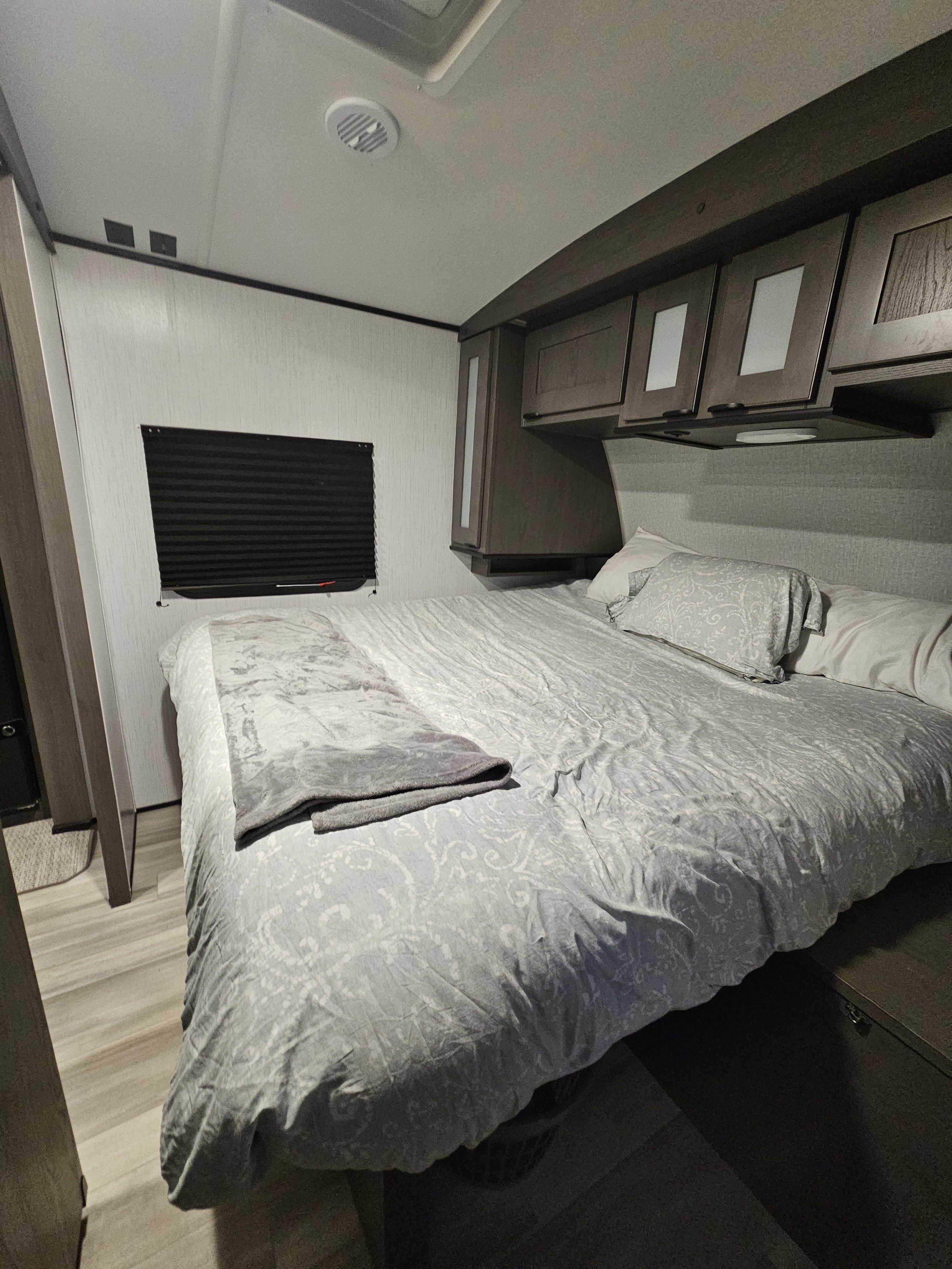 A neat and inviting bedroom inside an RV rental, featuring a comfortable bed, overhead cabinets, and a window with blinds.. Cruiser RV MPG 2840BH 2023