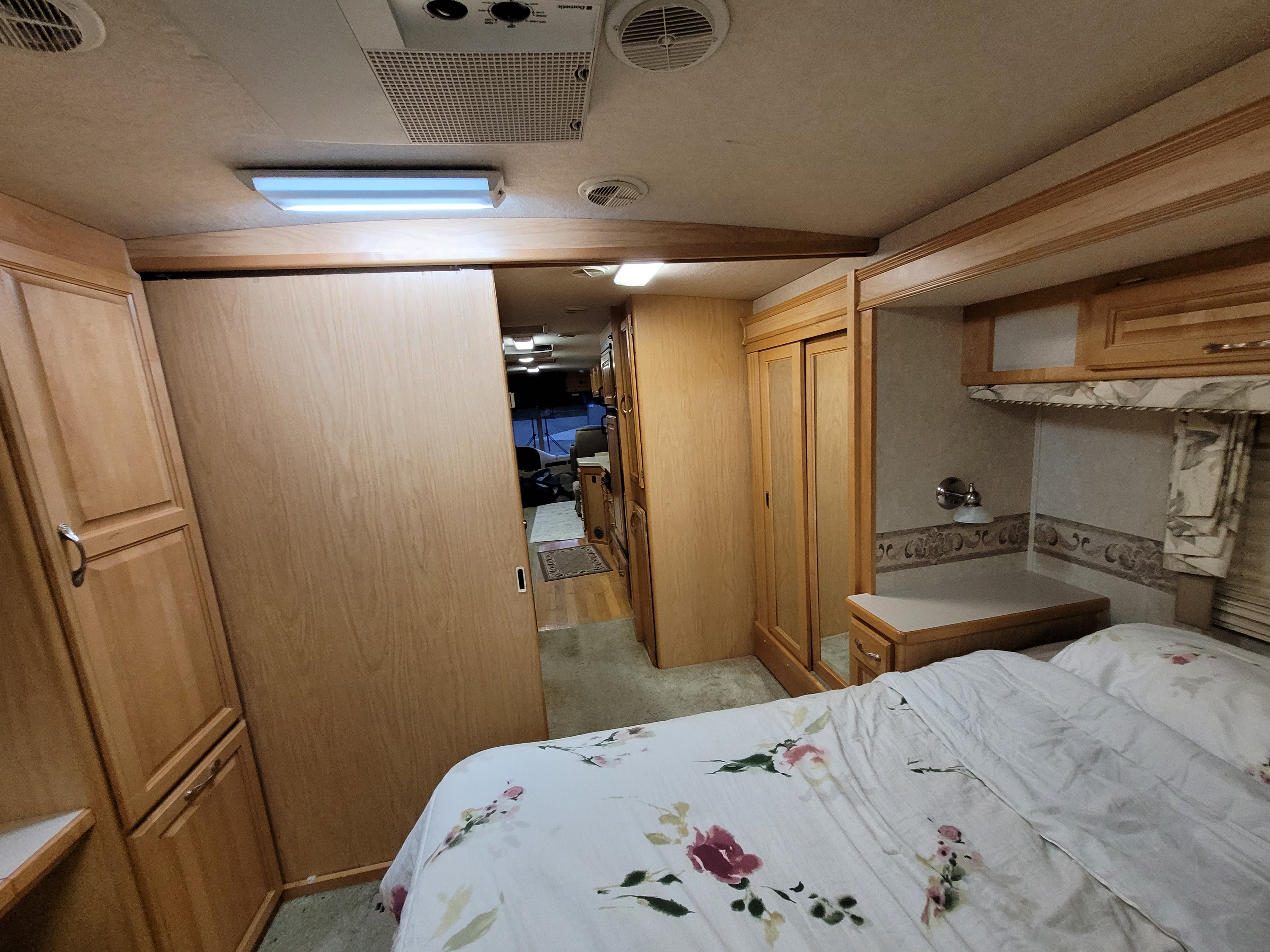 Inside a cozy RV rental bedroom looking towards the hallway, featuring floral bedding, wood cabinetry, and overhead AC unit.. Triple E Commander 2004
