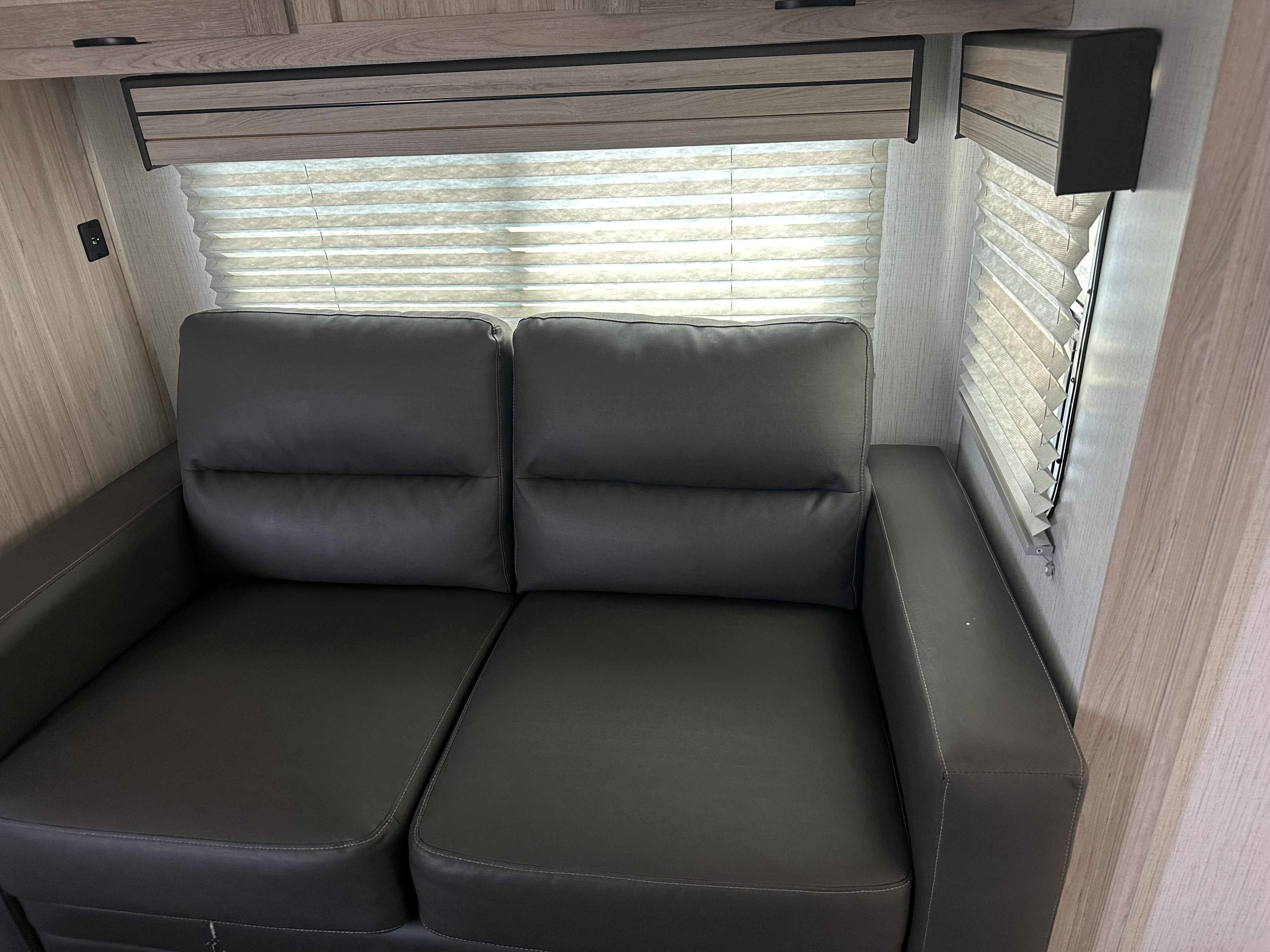 Modern RV rental living space featuring a dark gray loveseat, pleated window blinds, and light wood paneled walls.. EarthCruiser 248RKS 2022