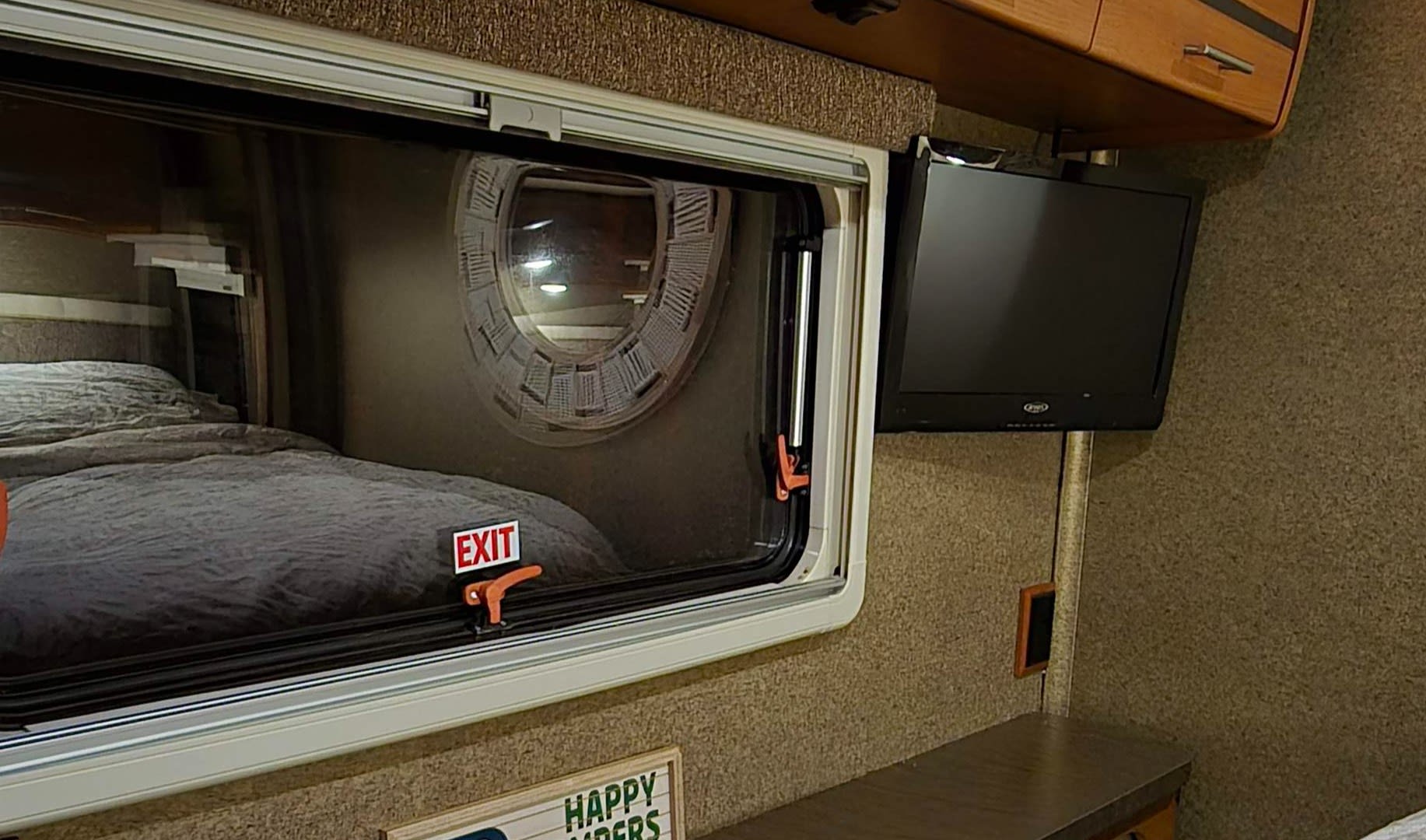 Inside an RV rental: a made bed by an exit window that reflects a unique round window. A small TV is mounted nearby.. Winnebago Via 2013