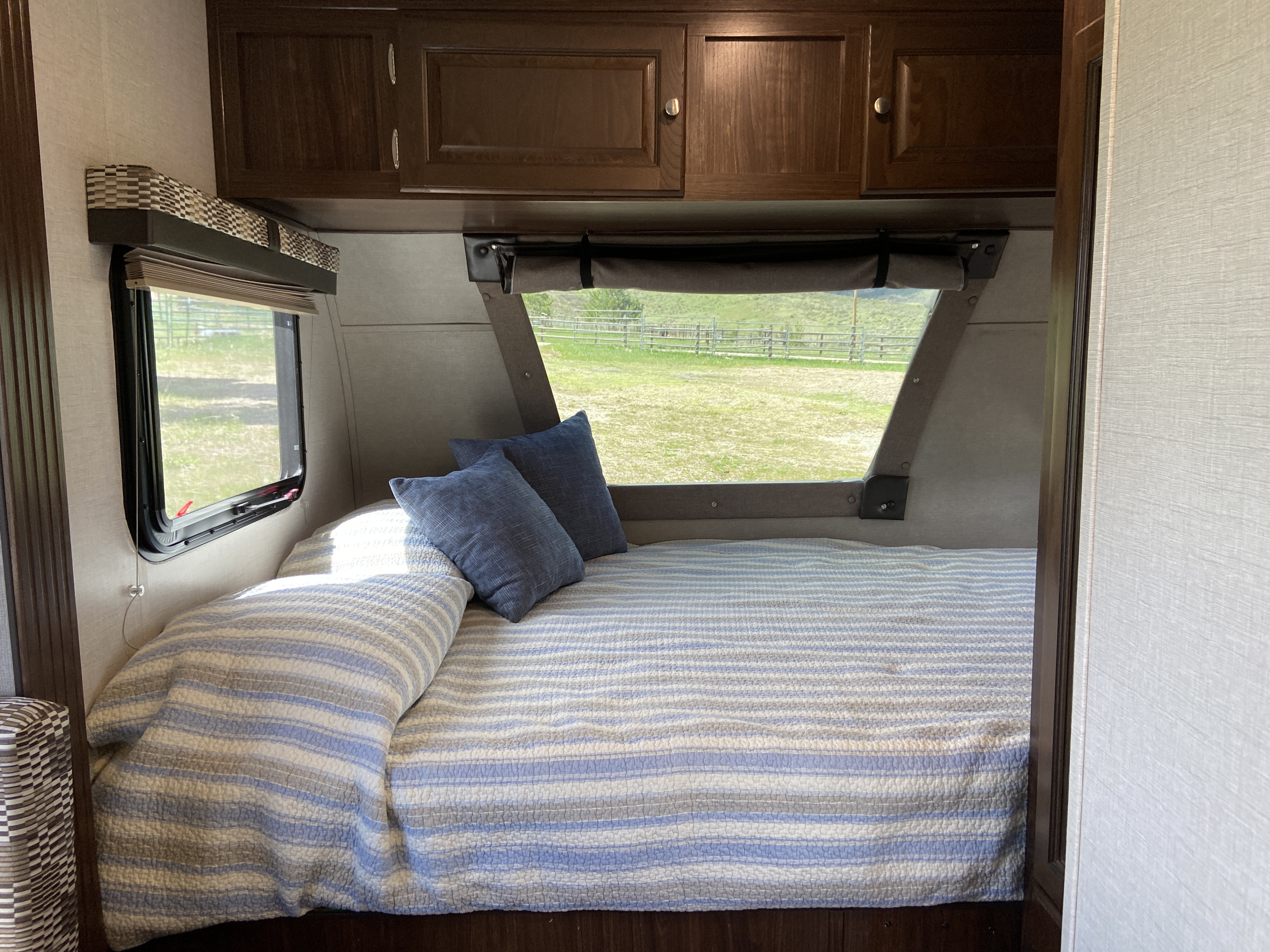 RV rental bedroom with striped bedding, blue pillows, overhead cabinets, and a window view of a grassy field.. Forest River Palomino Palomini 2018