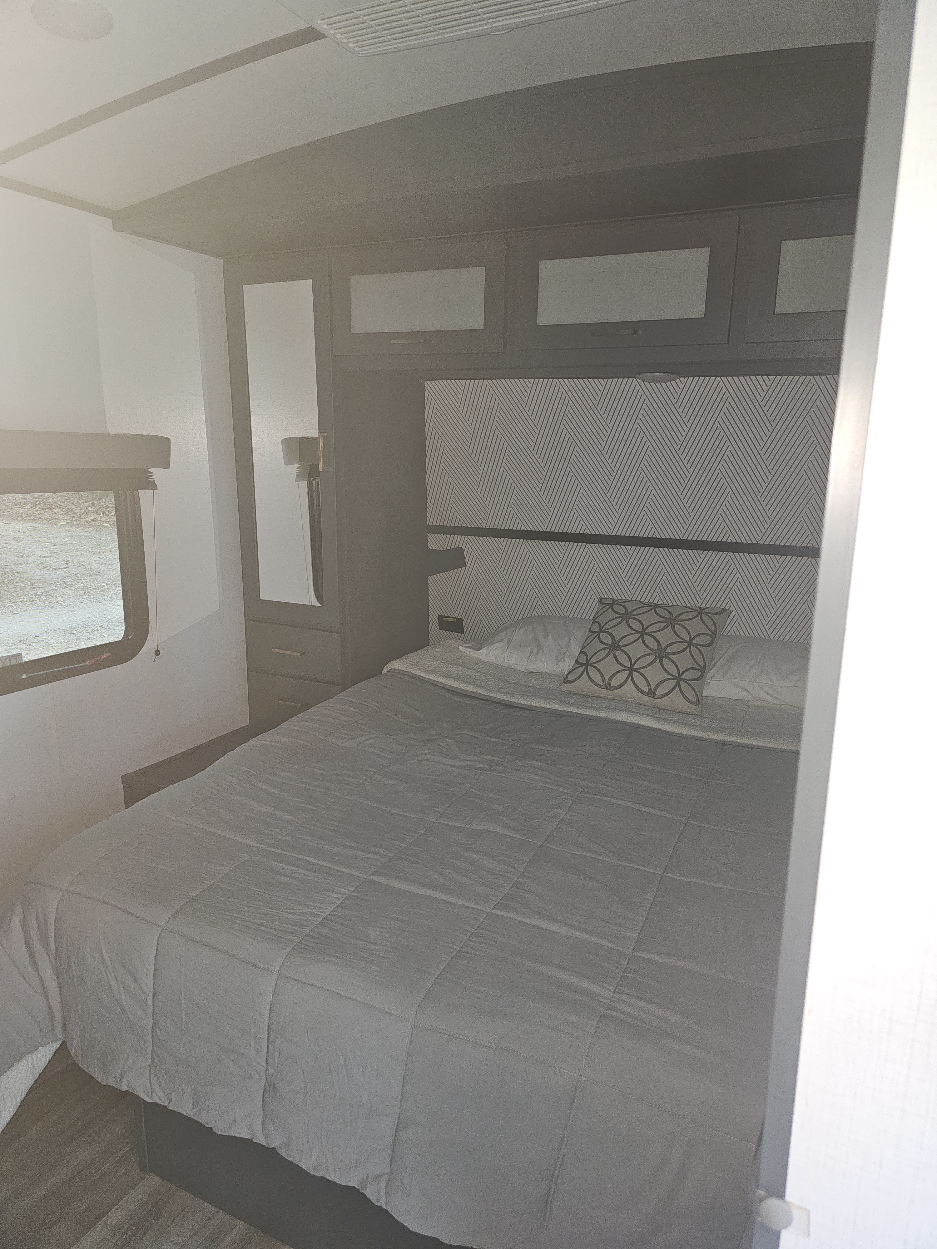 Modern RV rental bedroom with a comfy grey bed, patterned headboard, overhead cabinets, and a side window.. Keystone RV Bullet Ultra Lite 2022