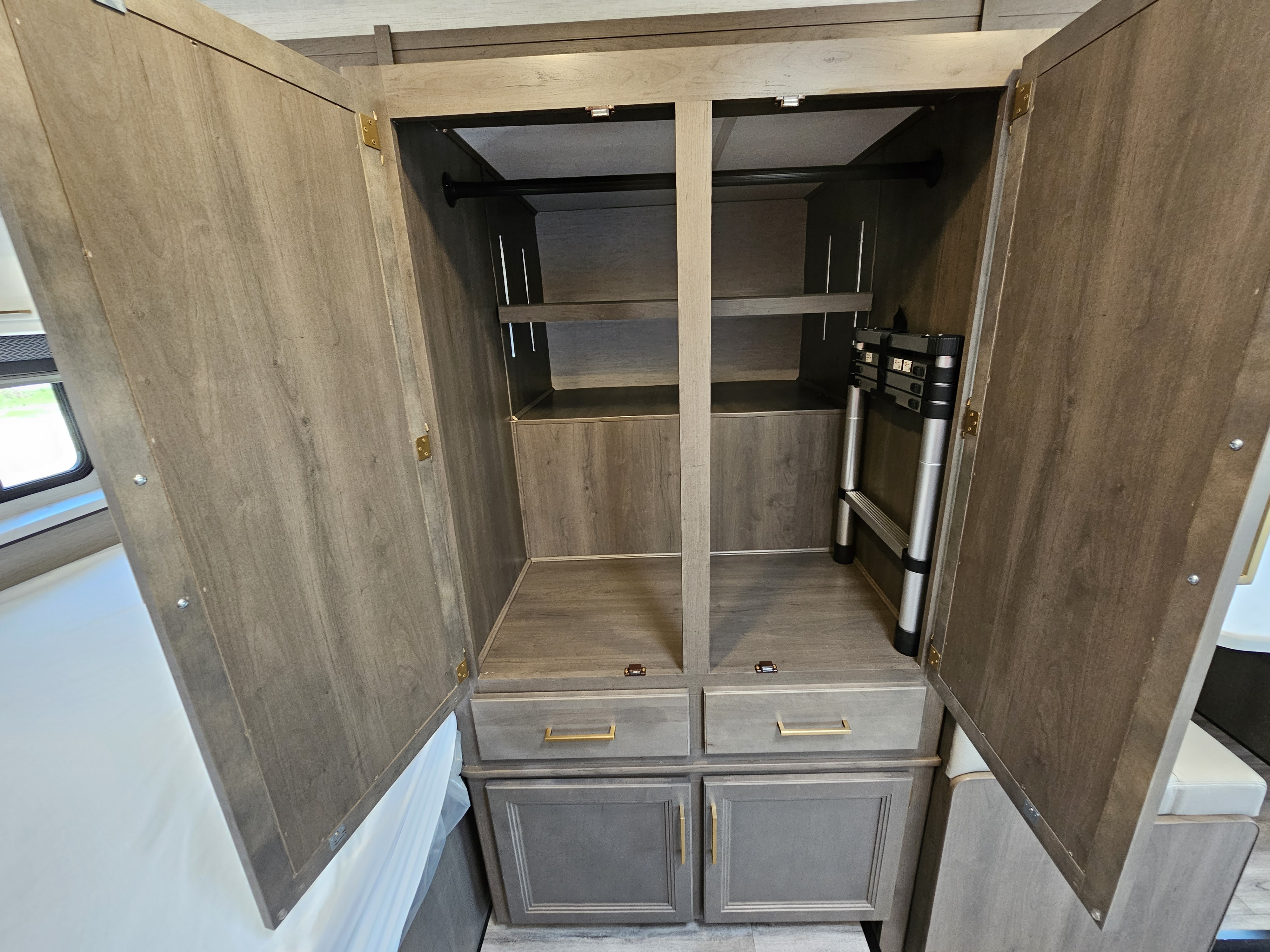 Open wooden closet in an RV rental, showing a hanging rod, adjustable shelving, drawers, and a stored ladder. Practical interior.. Thor Motor Coach Four Winds 2025