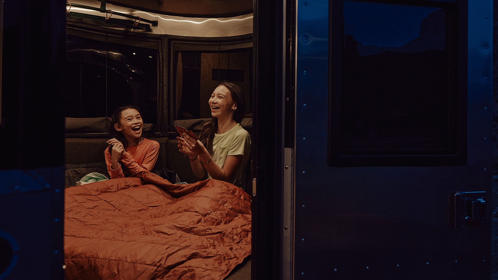 RV rental fun: Two girls, one in a sleeping bag, laughing & playing cards in a cozy camper at night.. Airstream Basecamp 2024