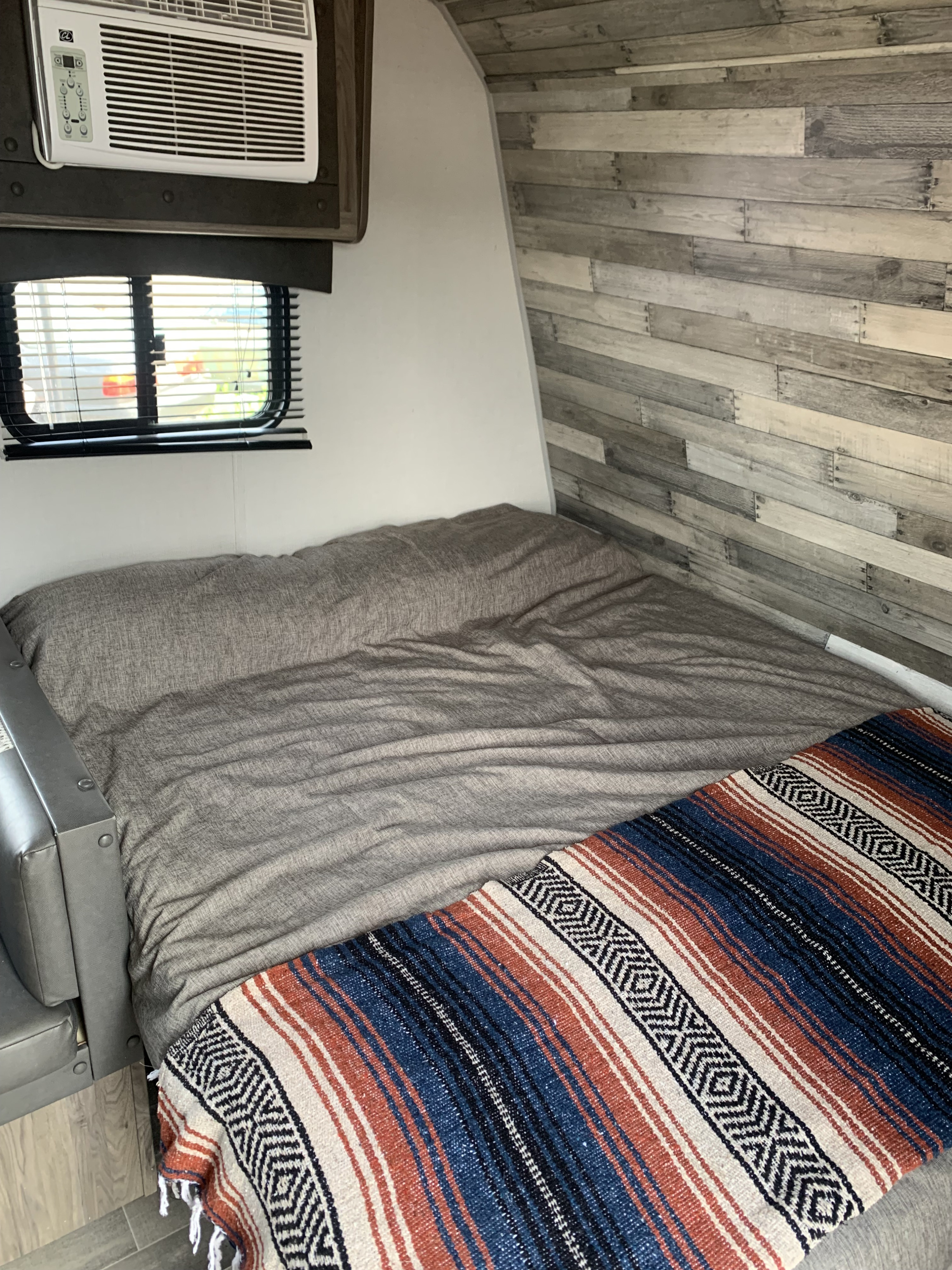 RV rental interior: cozy bedroom with grey bed, colorful striped blanket, window, AC unit, and wood panel wall.. Zinger BH 18 2021