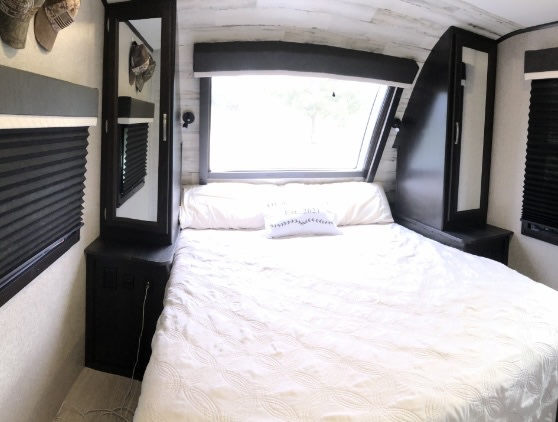 This RV rental bedroom features a queen bed with white bedding, a large front window, and dark built-in storage units on each side.. Jayco White Hawk 2021