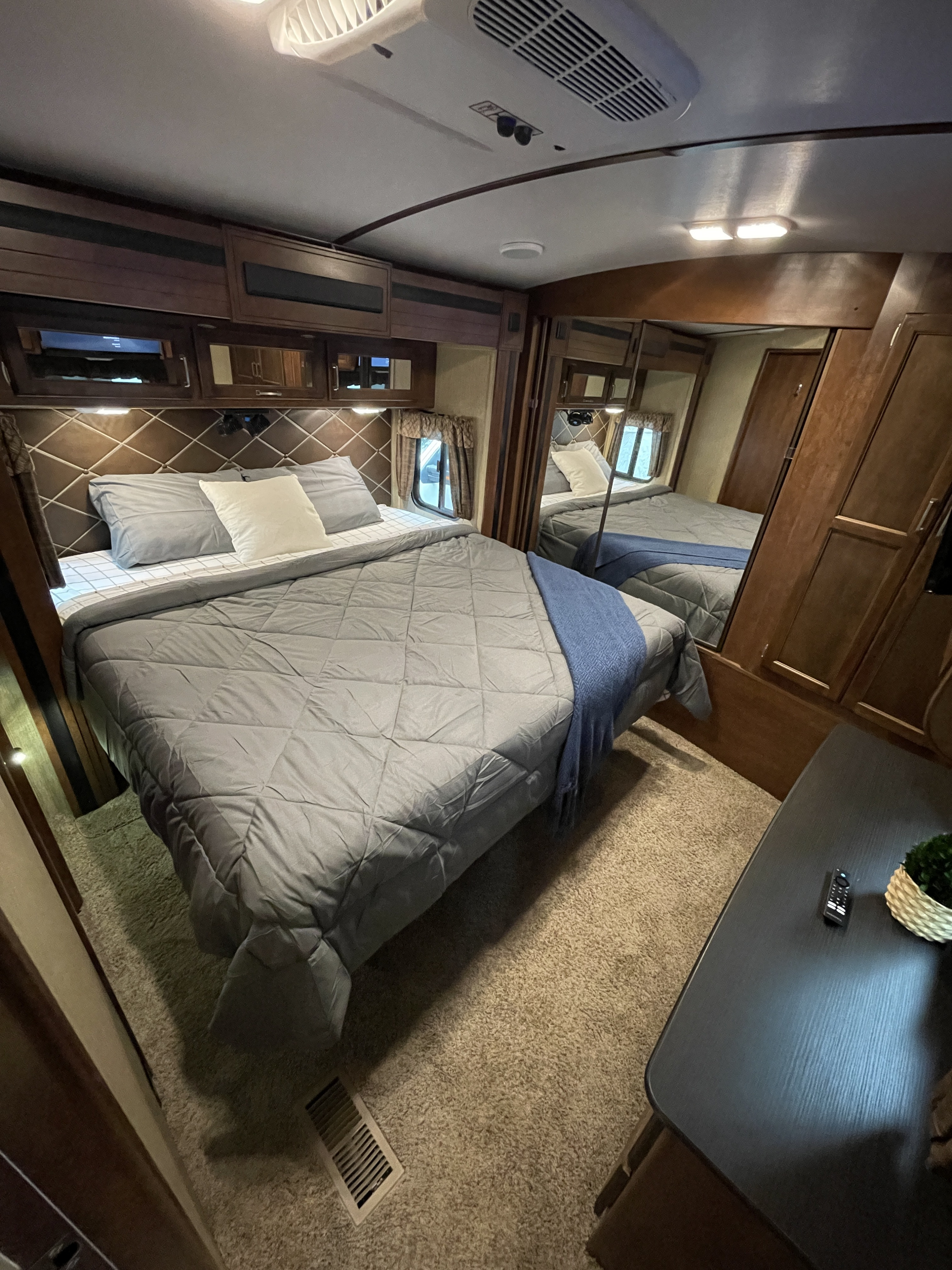 Cozy rv rental bedroom features a diamond-patterned headboard, grey bedding, overhead cabinets, and a mirror.. Keystone RV Outback Super-Lite 2018