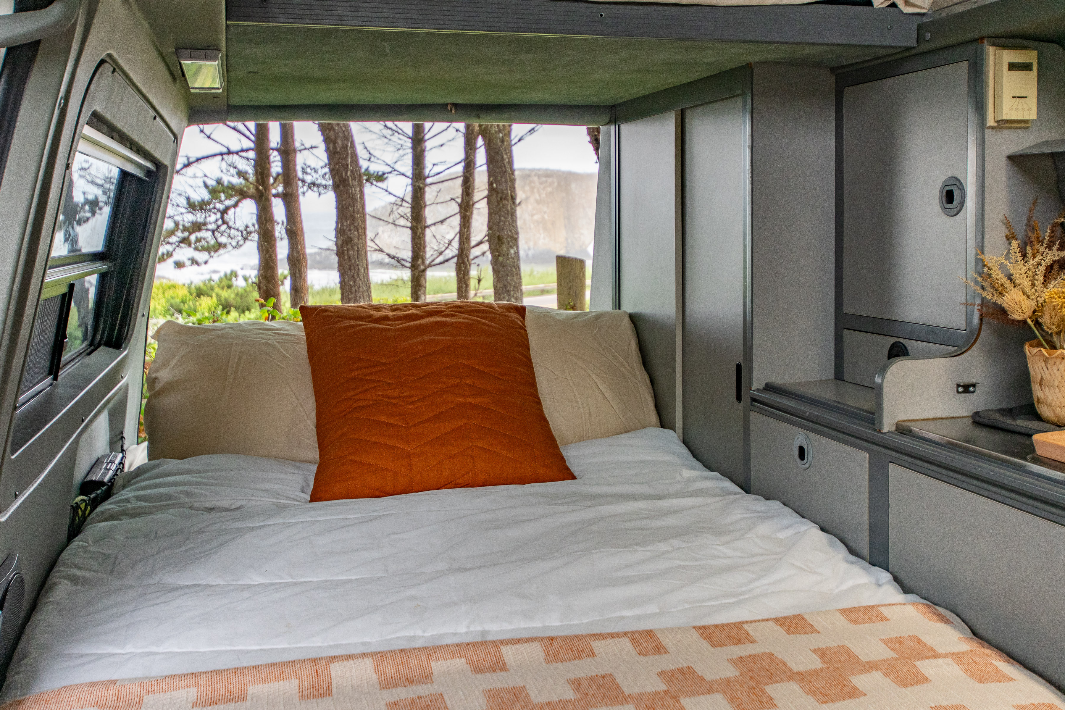 Inside an RV rental: a comfy bed with an ocean view through the window, and compact storage.. Volkswagen EuroVan 1996