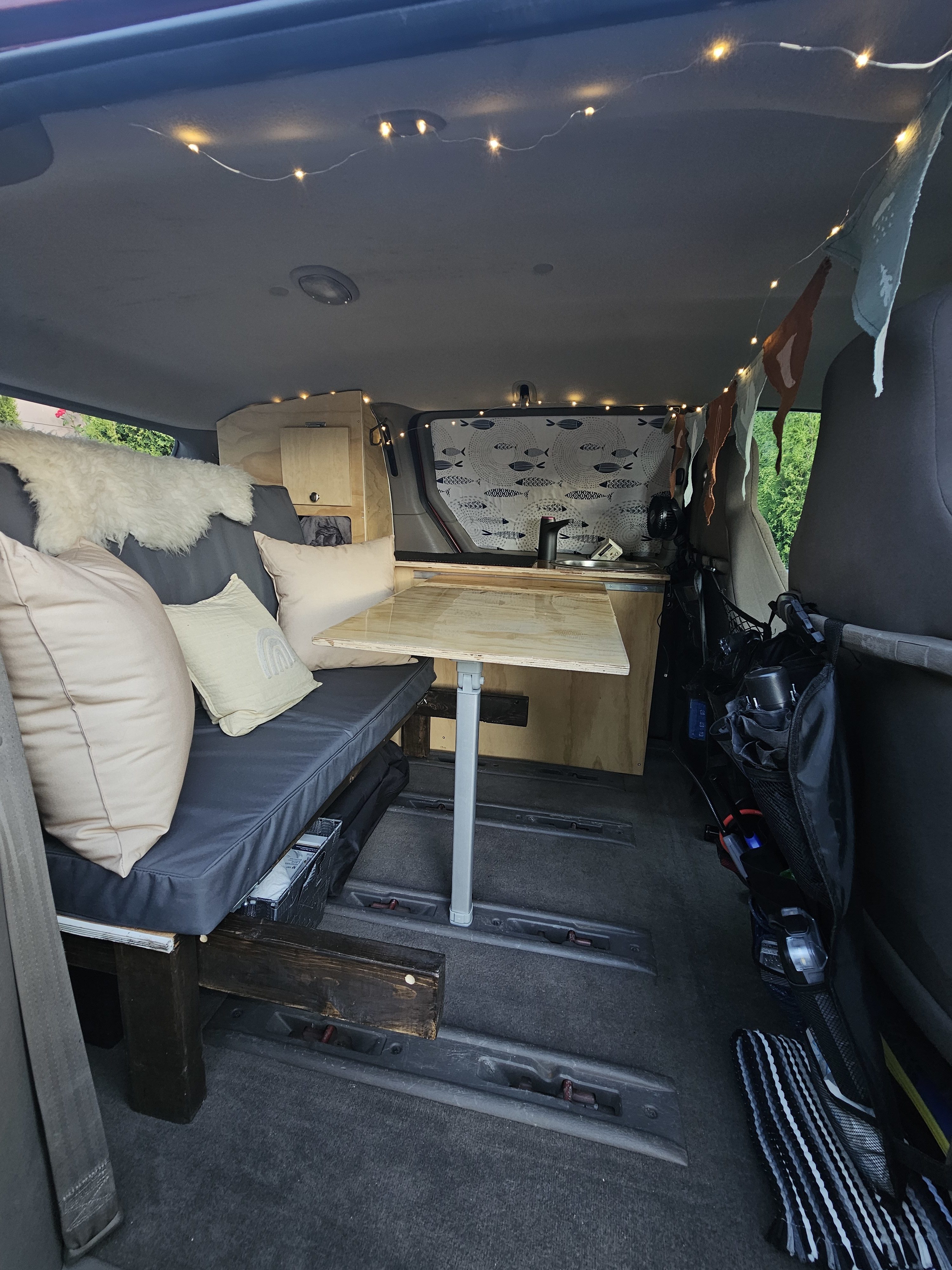 Inside a comfy RV rental van: a bench with cushions, portable table, wood cabinetry, and string lights create a cozy mobile home.. Dodge Caravan 2003